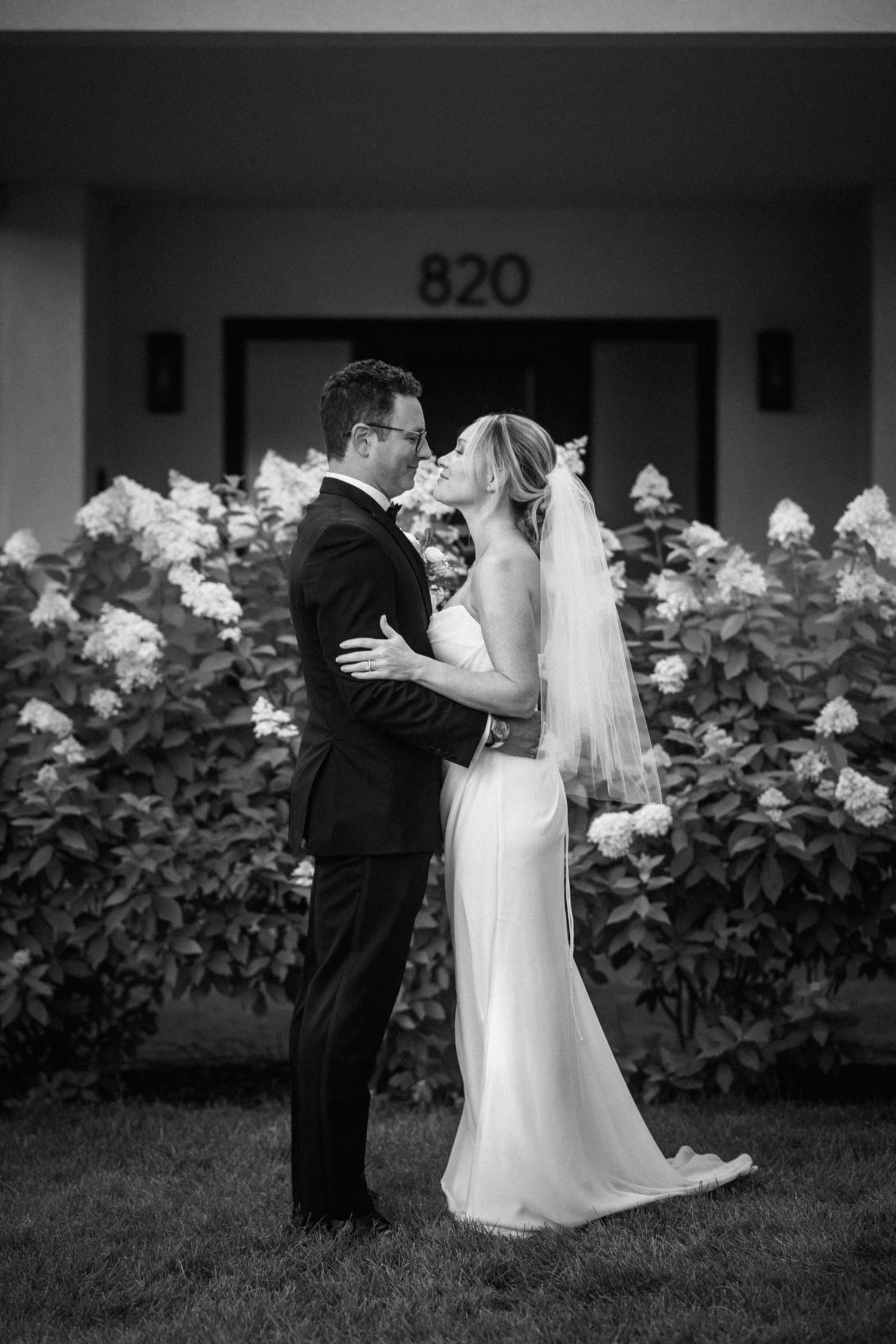 Black and white photo of bride and groom sharing a kiss in front of blooming hydrangeas outside building 820.