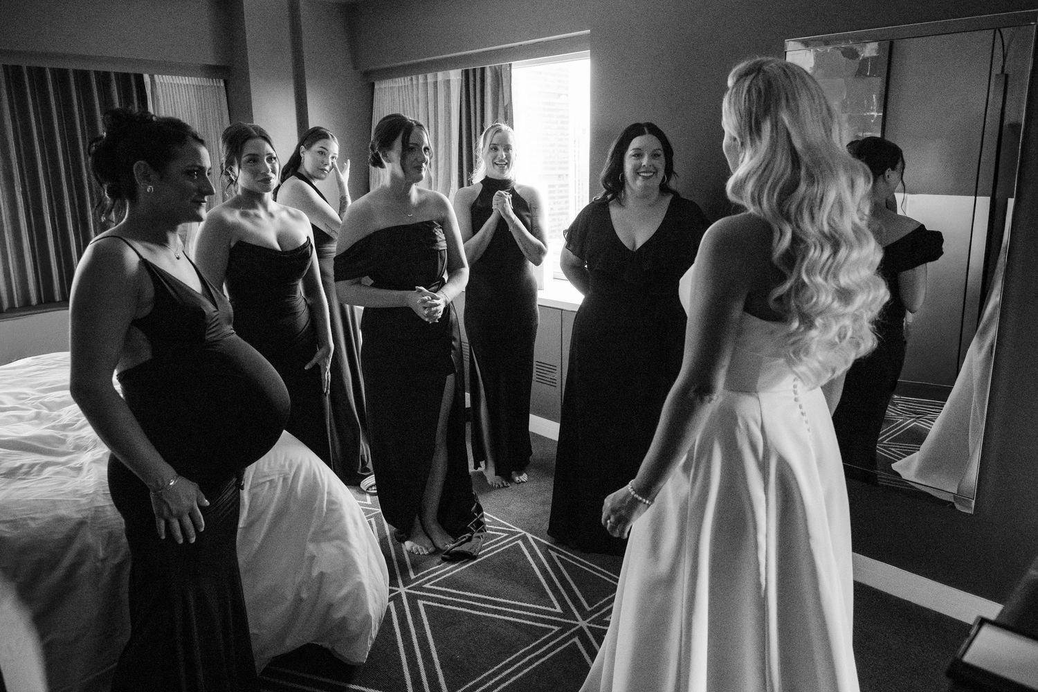 Black and white photo of bride in white gown doing first look reveal with bridesmaids in black dresses in hotel room.