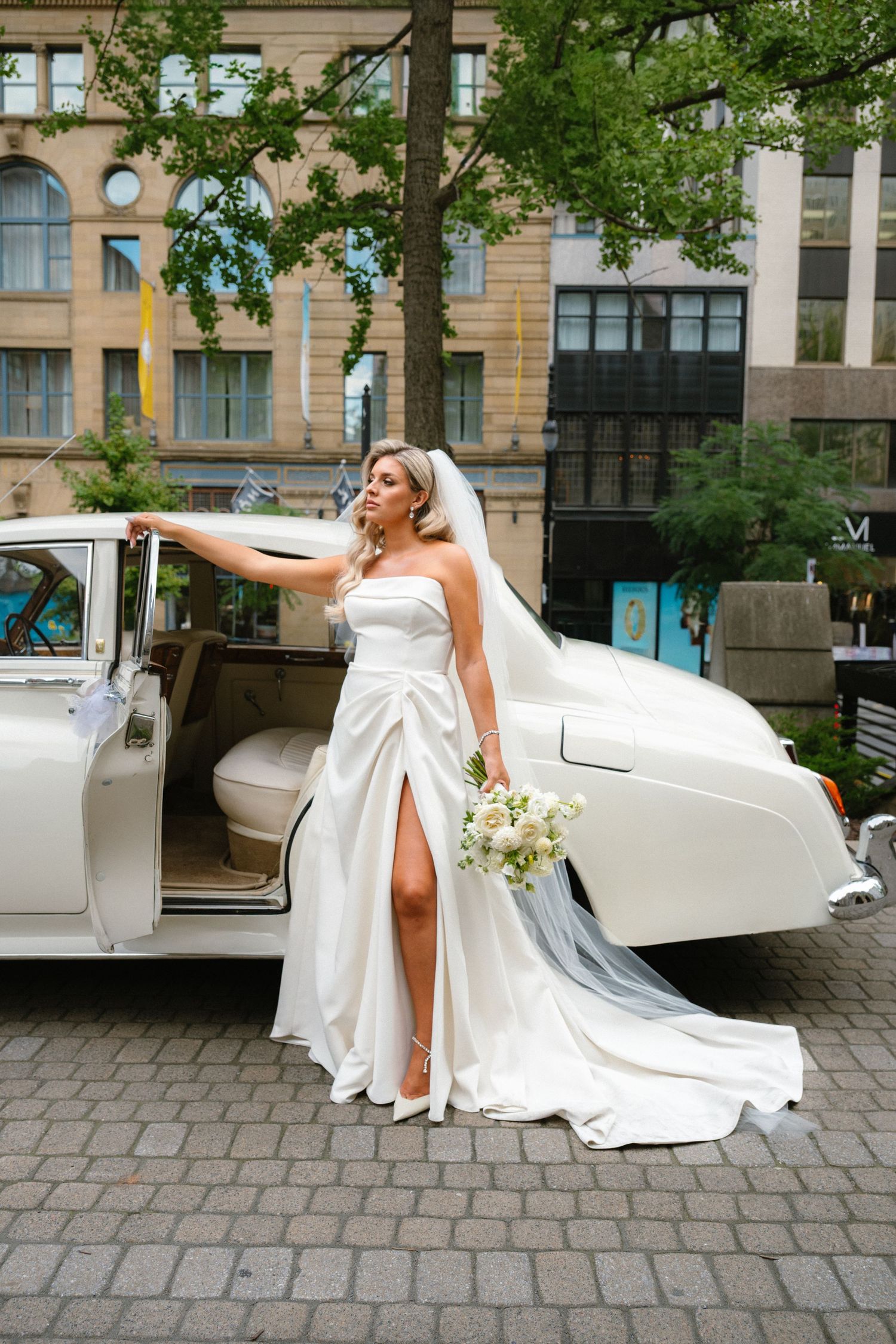 Beautiful bride in strapless white wedding gown with veil holding bouquet beside vintage white car outdoors.