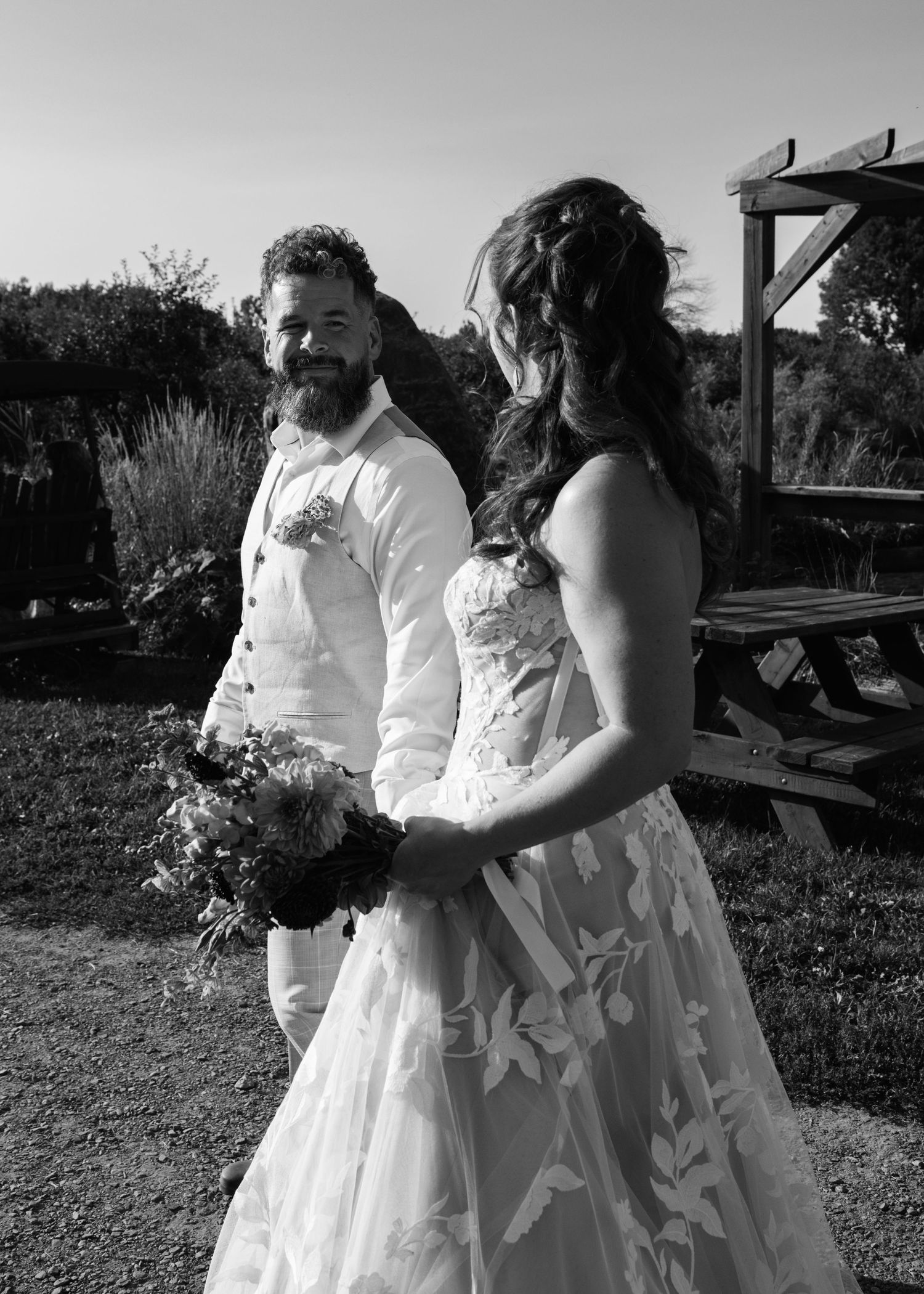 Bearded groom in white suit smiles at bride holding bouquet in black and white outdoor wedding photo.