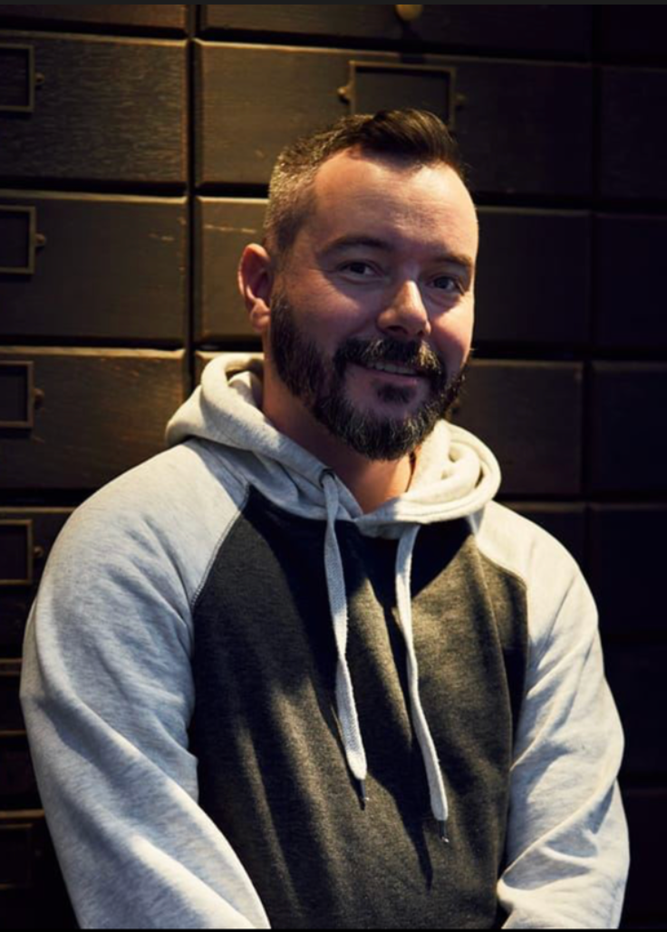 Smiling bearded man in gray hoodie posing against dark cabinet background.