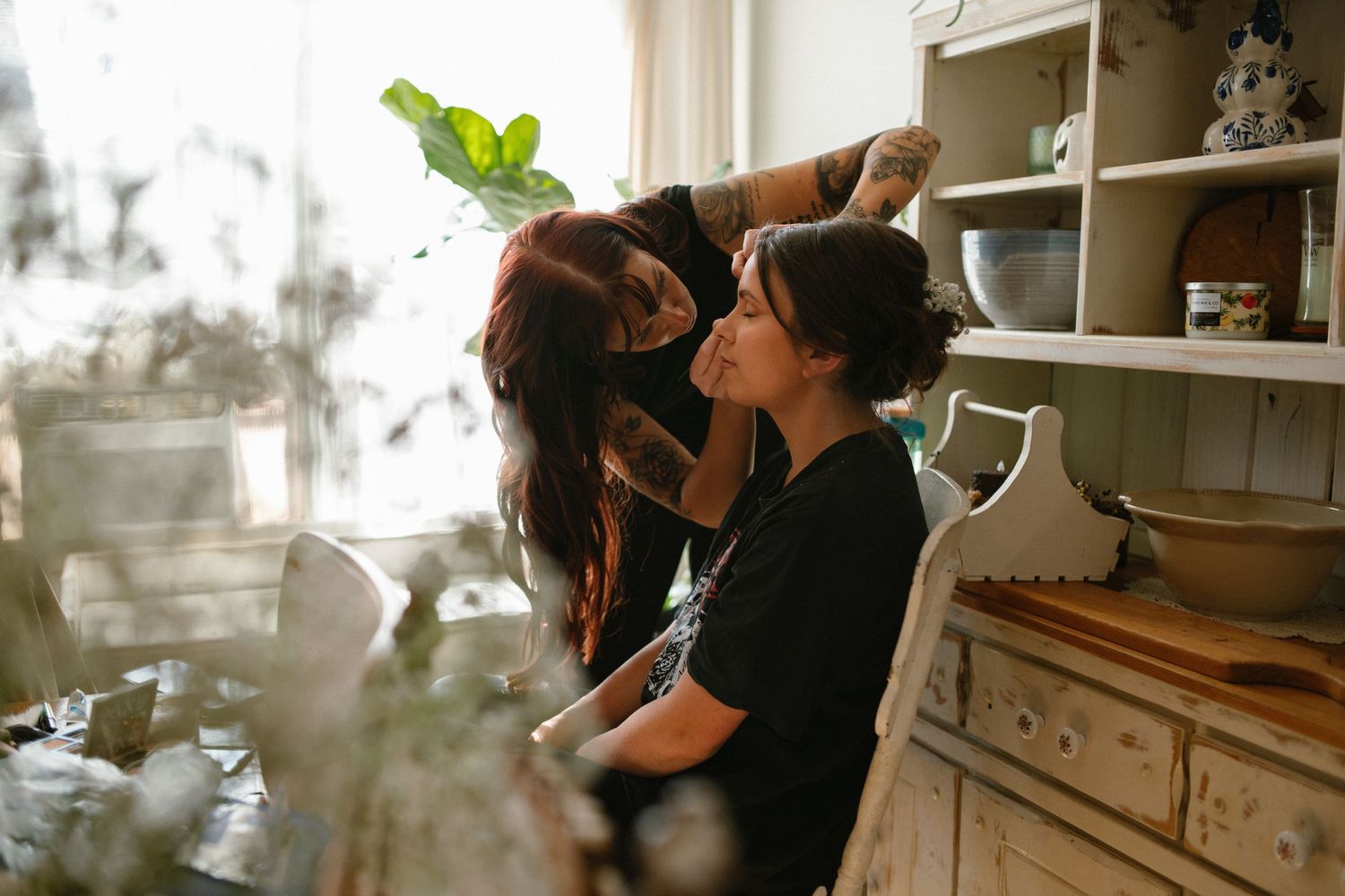 A makeup artist applies makeup to a woman seated near a rustic wooden dresser in a warmly lit room.