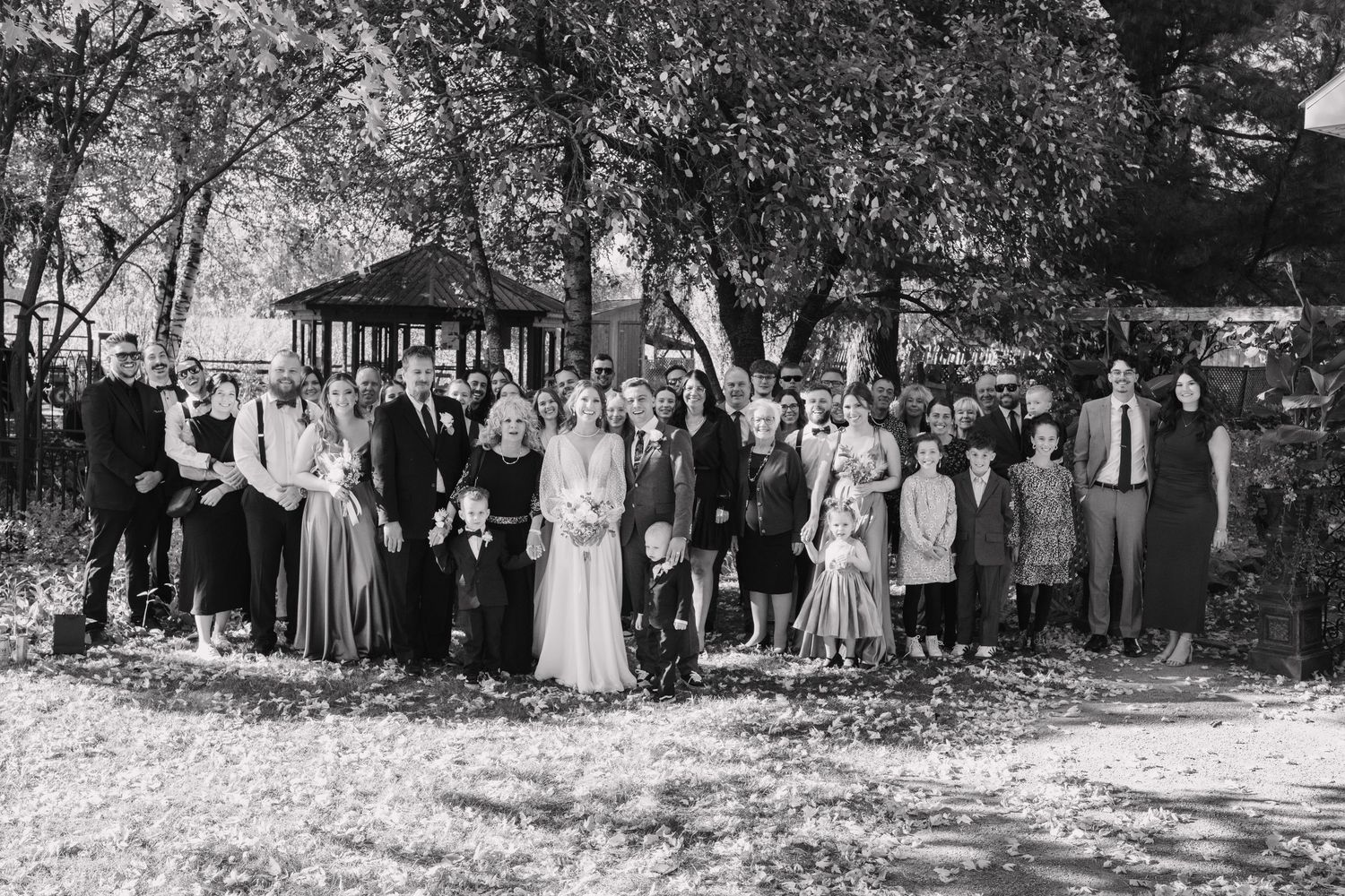 Black and white group wedding portrait of family and guests gathered near a park gazebo outdoors.