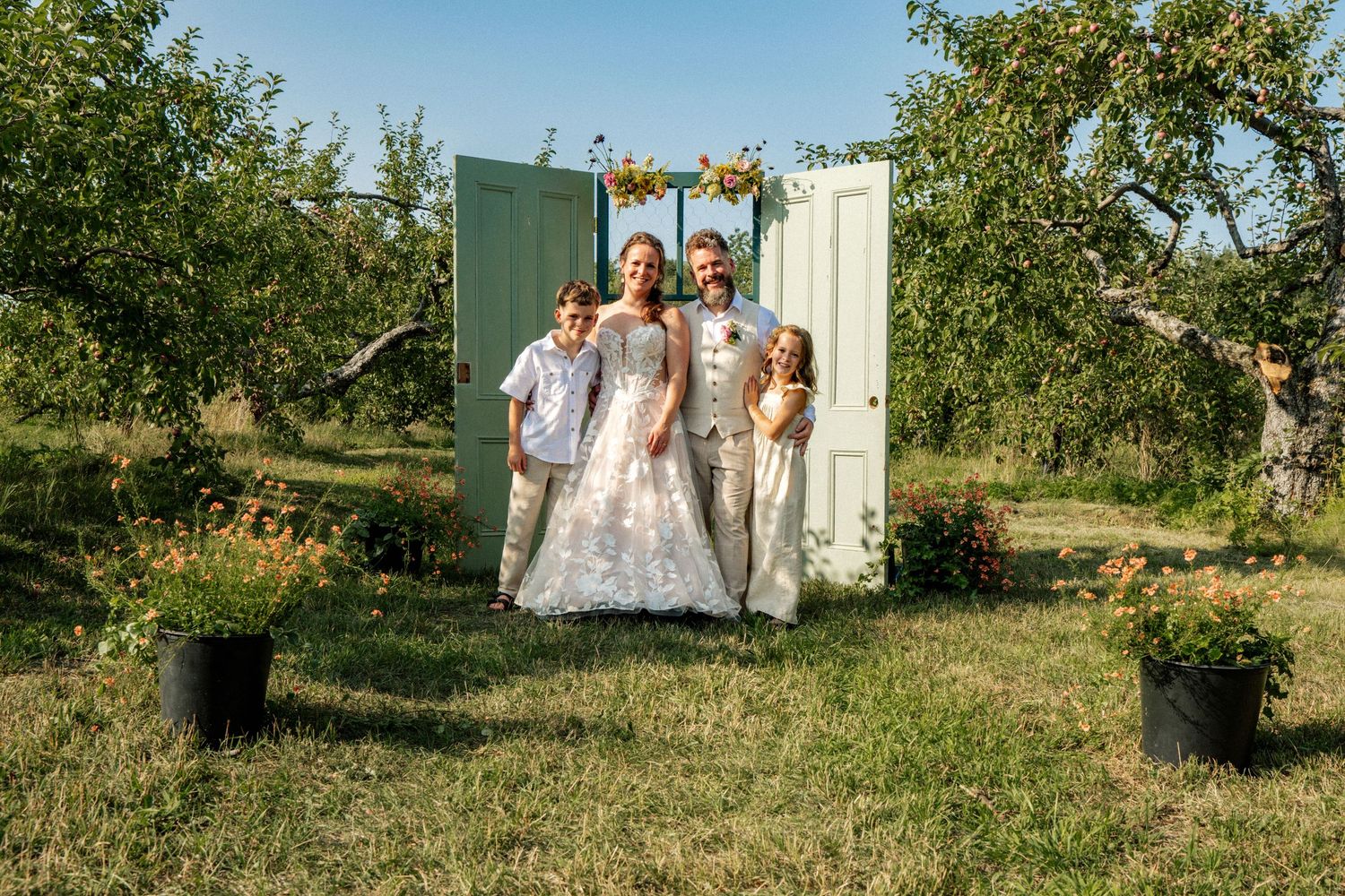 Happy wedding family portrait with bride, groom, and two children in an apple orchard.