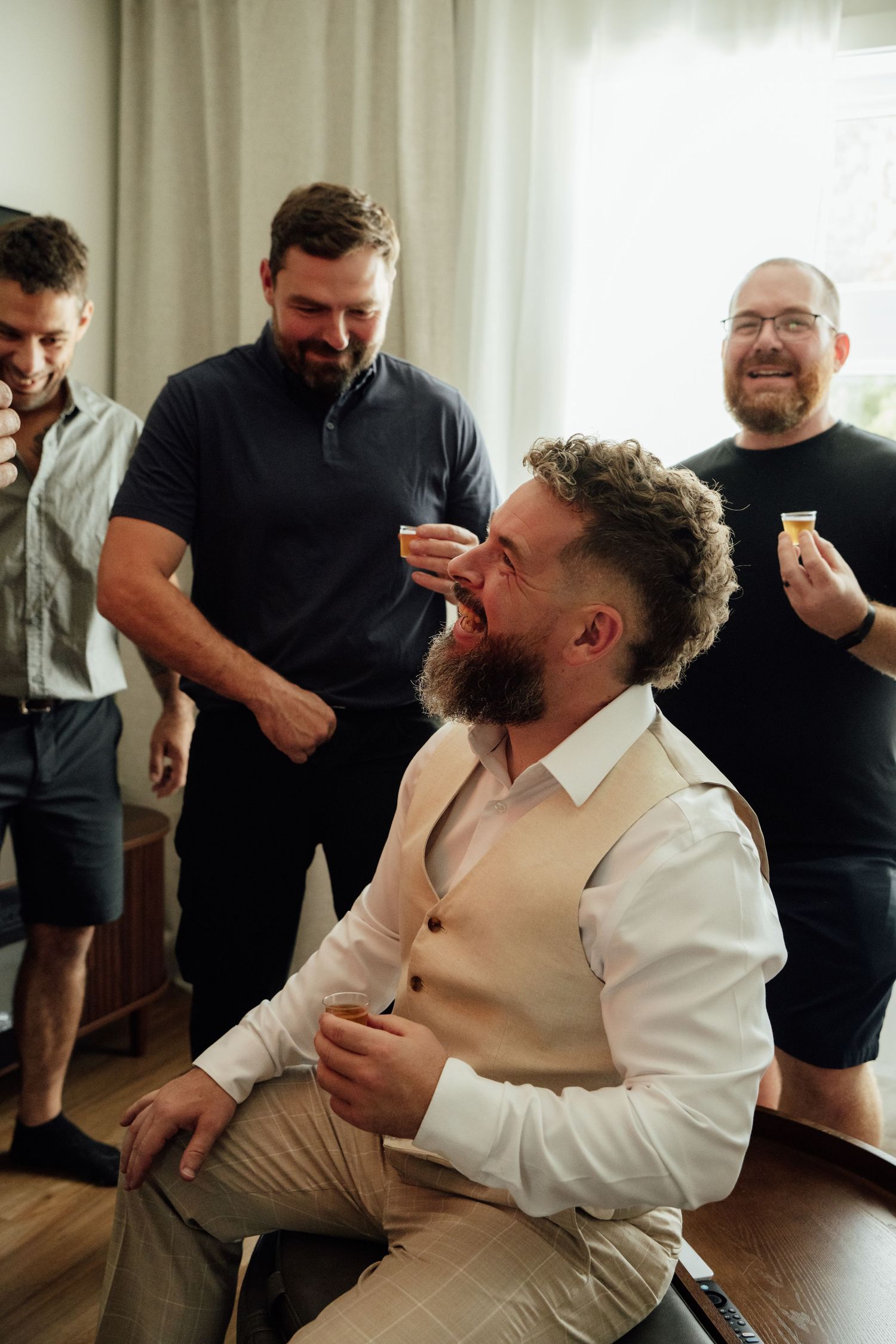 Groom laughing with groomsmen during wedding preparations indoors.