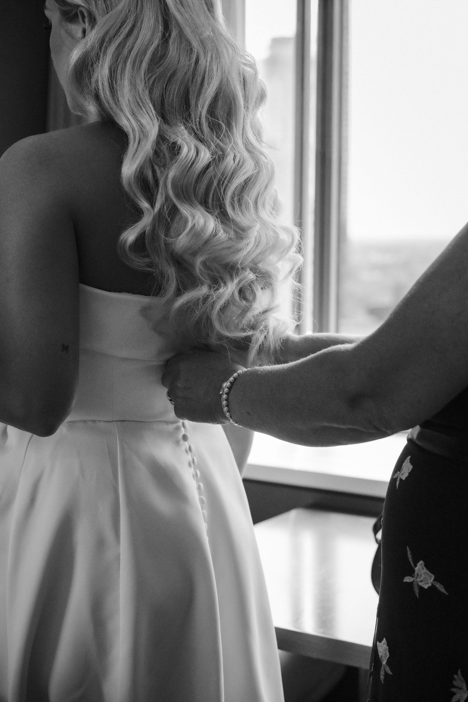 Black and white photo of bride having wedding dress buttoned up from behind near a bright window.