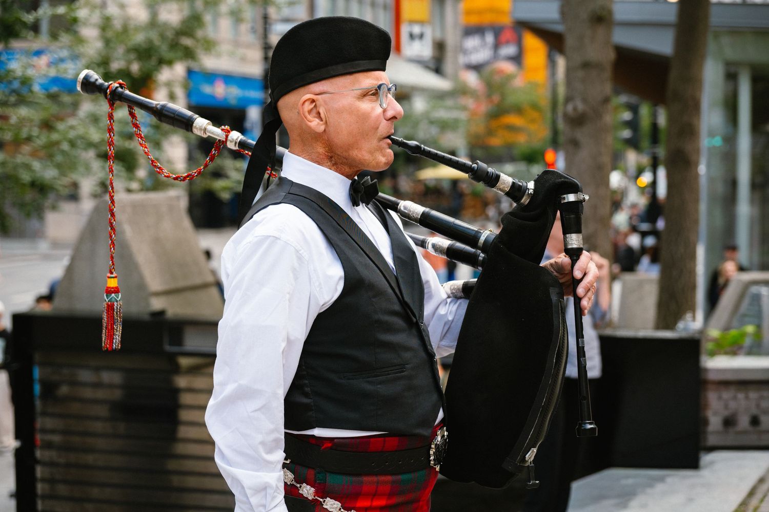 Scottish bagpiper in traditional tartan kilt performing outdoors on city street.