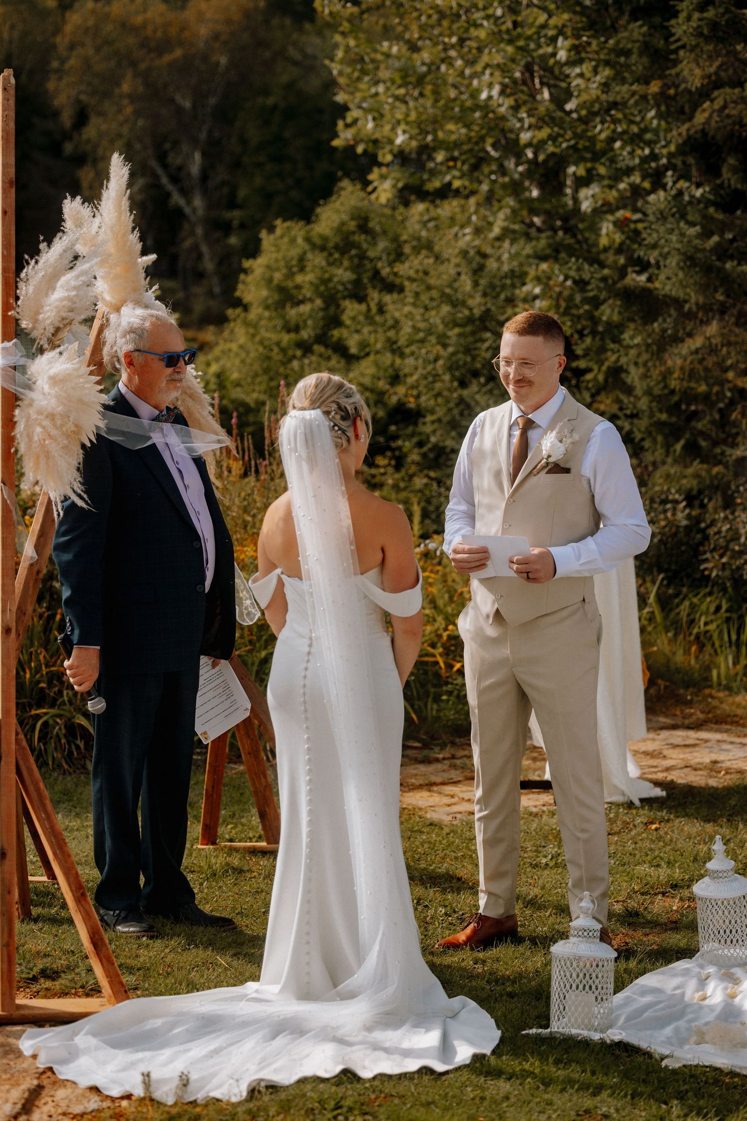Outdoor wedding ceremony with bride in white gown and groom in tan suit exchanging vows before an officiant.