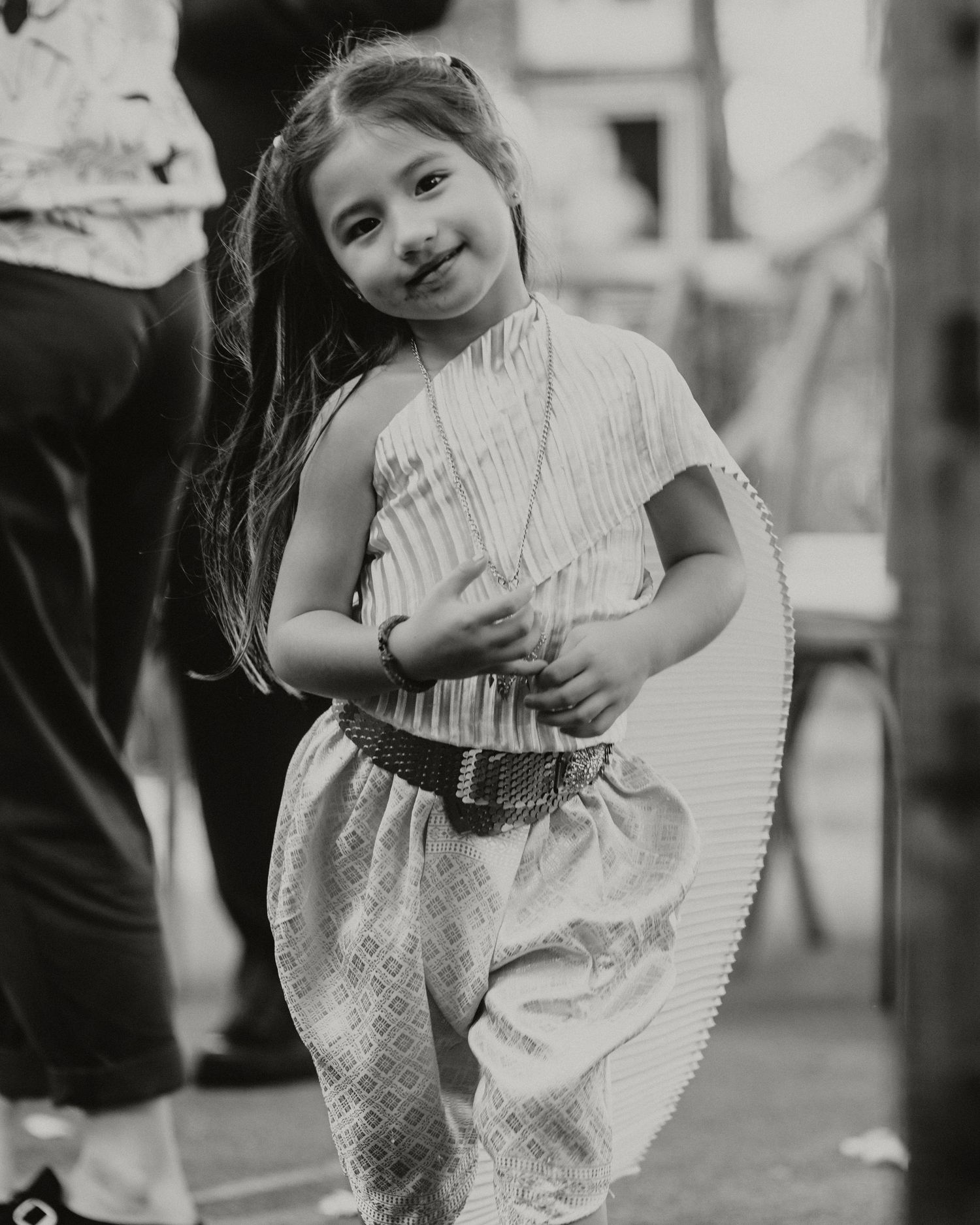 A smiling little girl in traditional cultural attire with a woven belt poses playfully in a black and white photo.