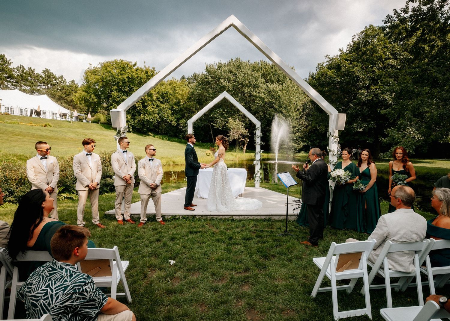 Outdoor wedding ceremony under geometric arch with bridal party on lush green lawn.