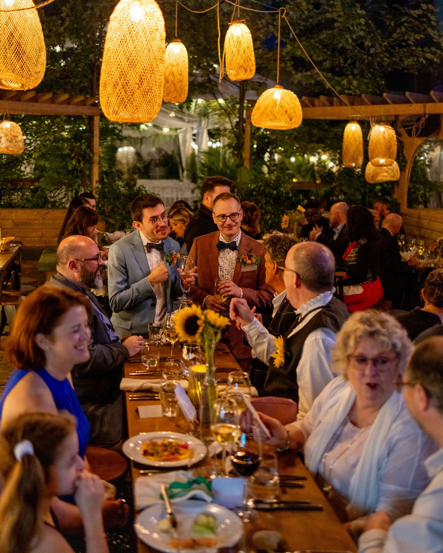 Guests dining at an elegant outdoor evening event with warm wicker pendant lights and sunflower centerpieces.
