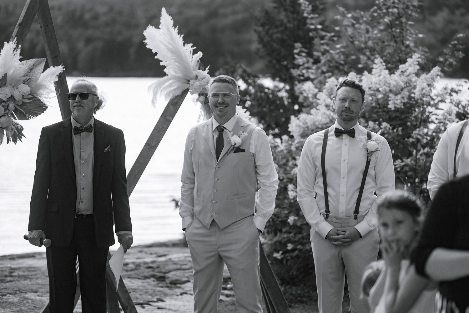 Black and white outdoor wedding ceremony with groom in light suit, groomsmen in bow ties, by a lakeside backdrop.