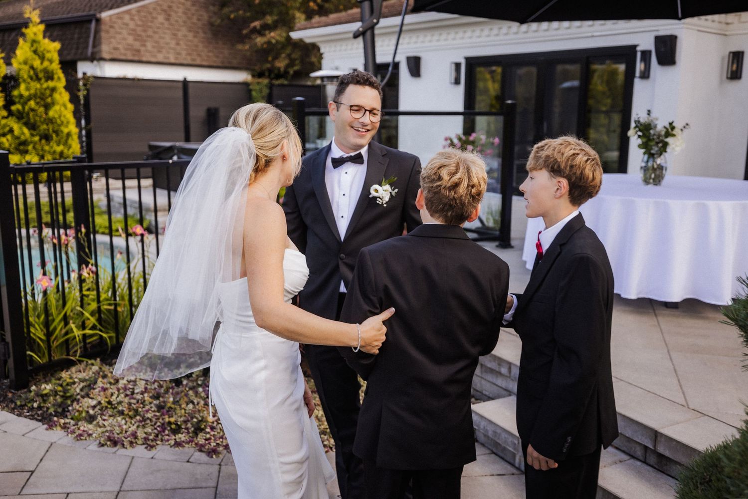 Bride and groom in tuxedo greet two young boys in suits after outdoor wedding ceremony.