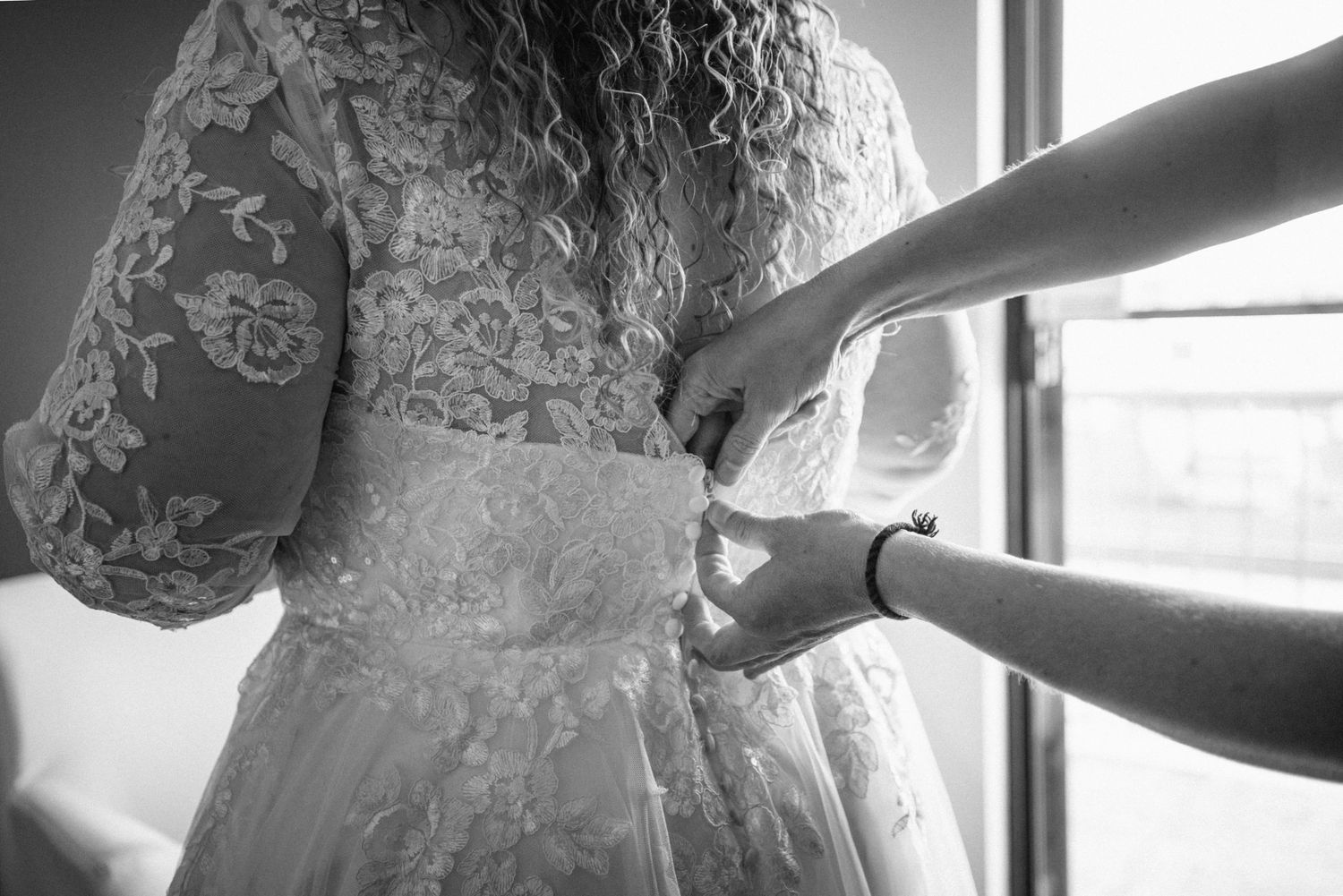 Black and white photo of a bride having her lace wedding dress buttoned up by a helper near a bright window.