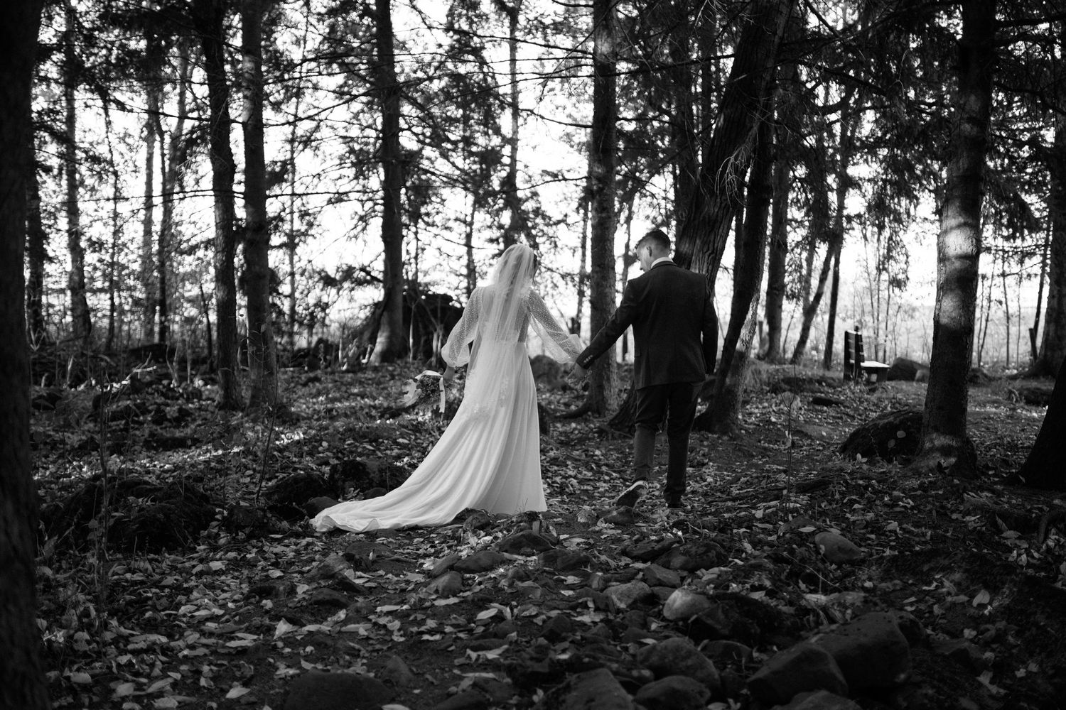 Black and white photo of bride and groom walking hand in hand through a wooded forest path.