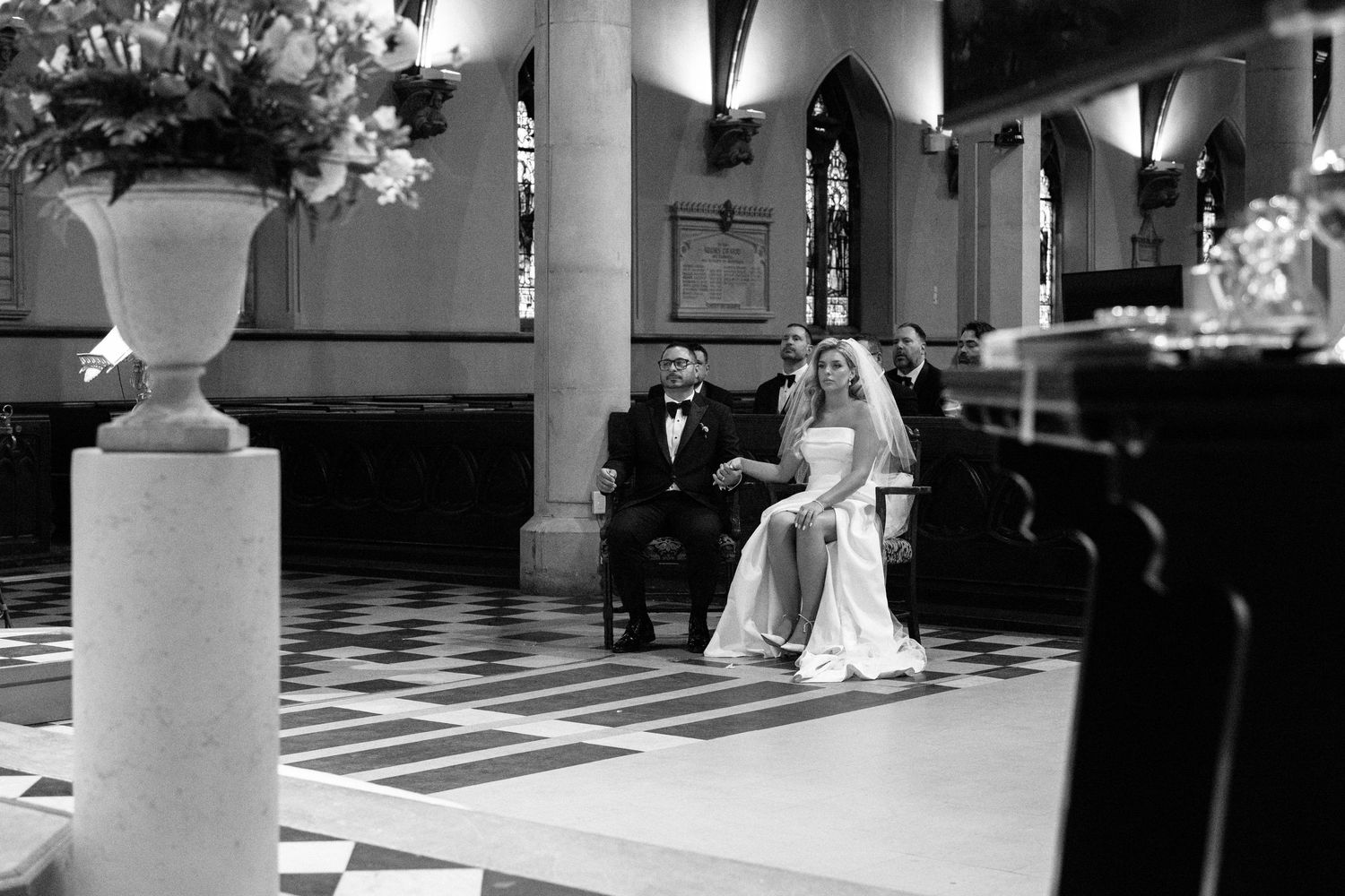Black and white photo of bride and groom seated during wedding ceremony inside elegant stone church.