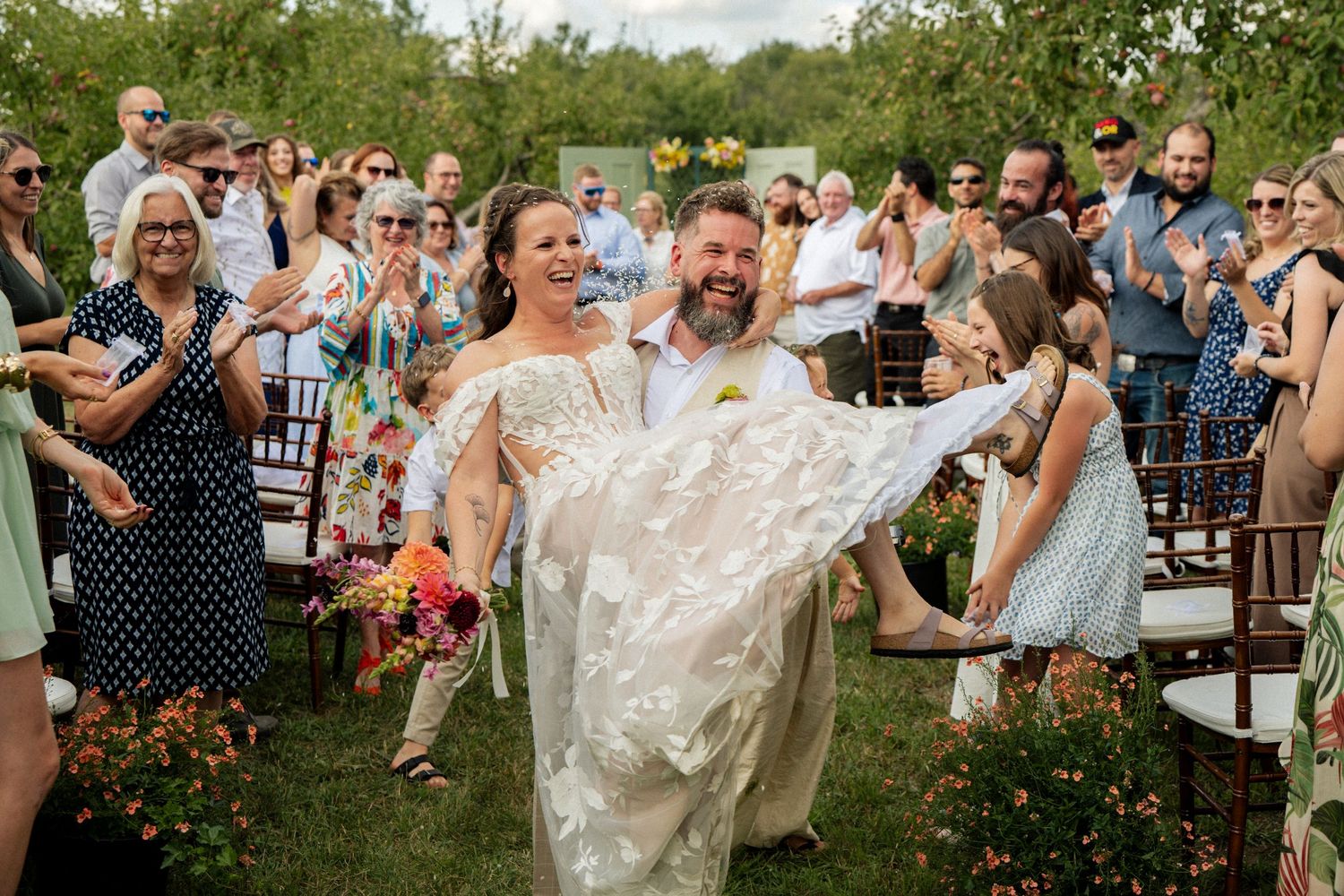 Groom carries laughing bride down the aisle as wedding guests cheer at an outdoor orchard ceremony.