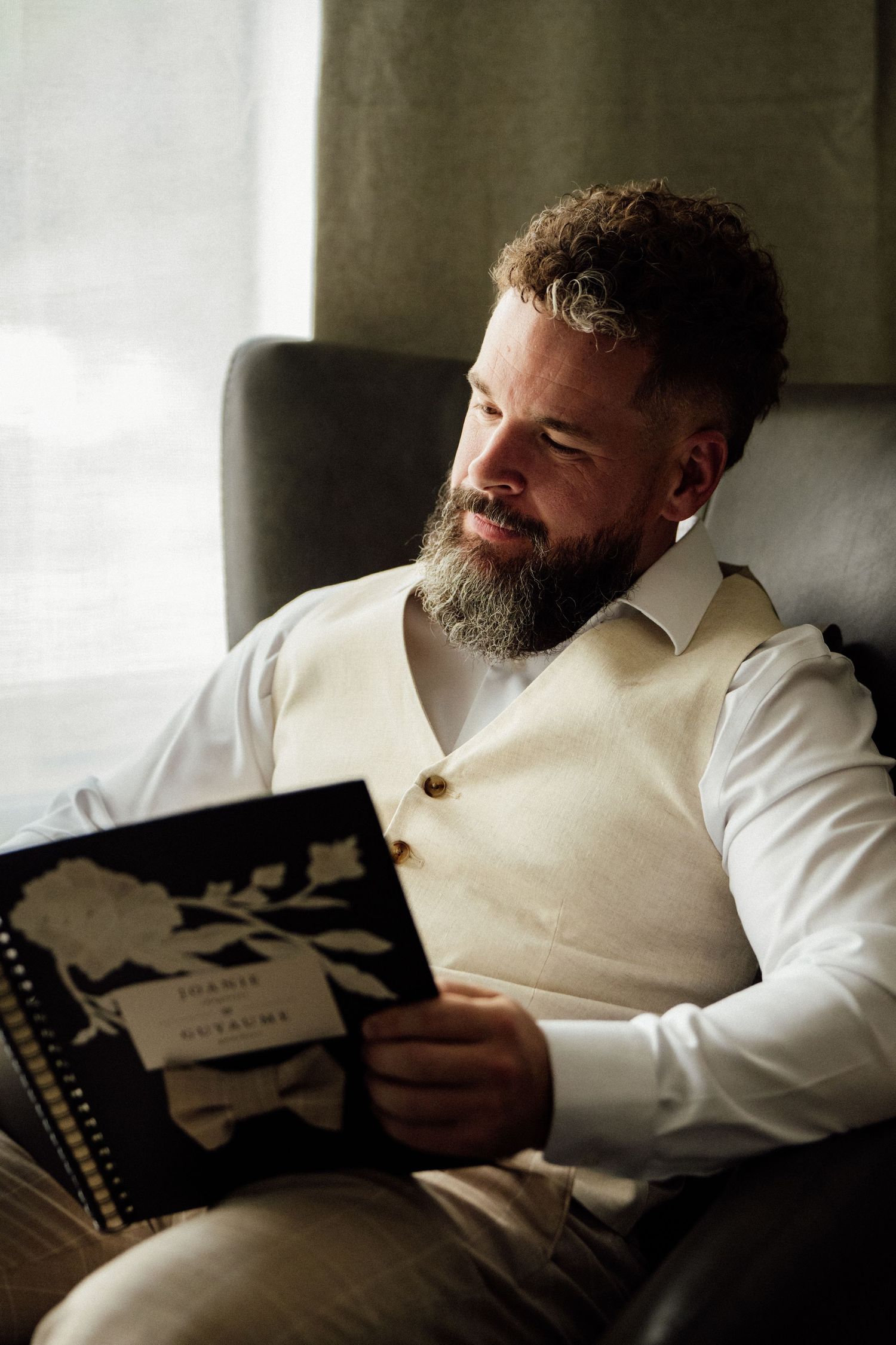 Bearded groom in white shirt and cream vest reads wedding program while seated in a gray chair.