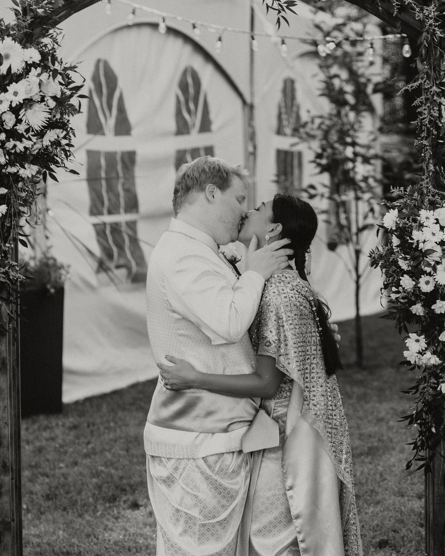 Black and white photo of a couple sharing a first kiss under a floral arch at an outdoor wedding ceremony.