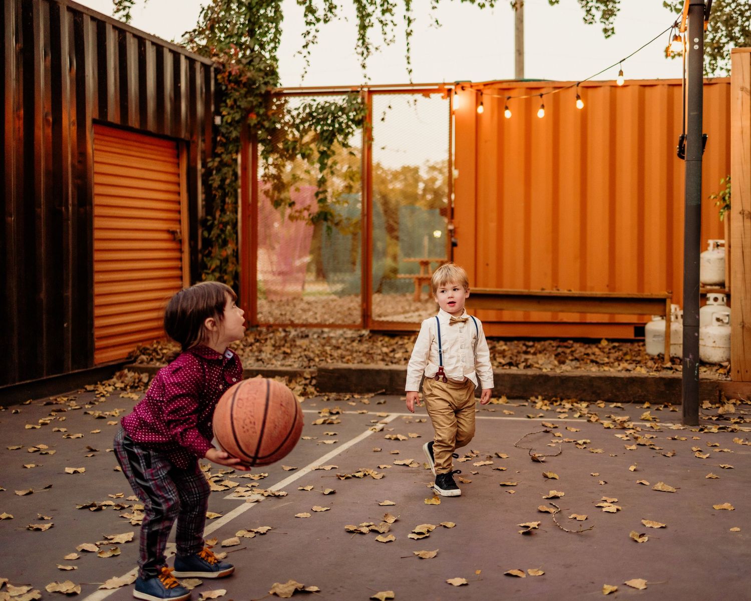 Two young children playing basketball outdoors on a court surrounded by orange shipping containers and string lights.
