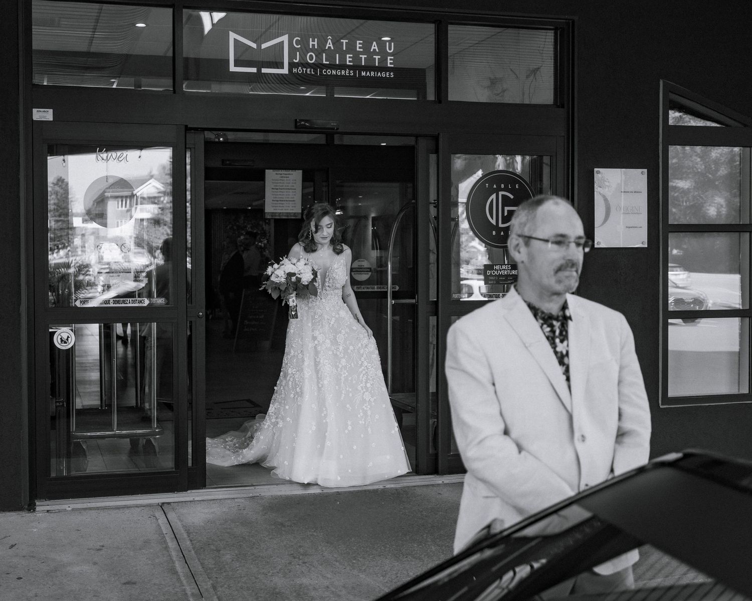 Black and white photo of a bride in a wedding gown exiting Chateau Joliette as a man in a white suit stands outside.