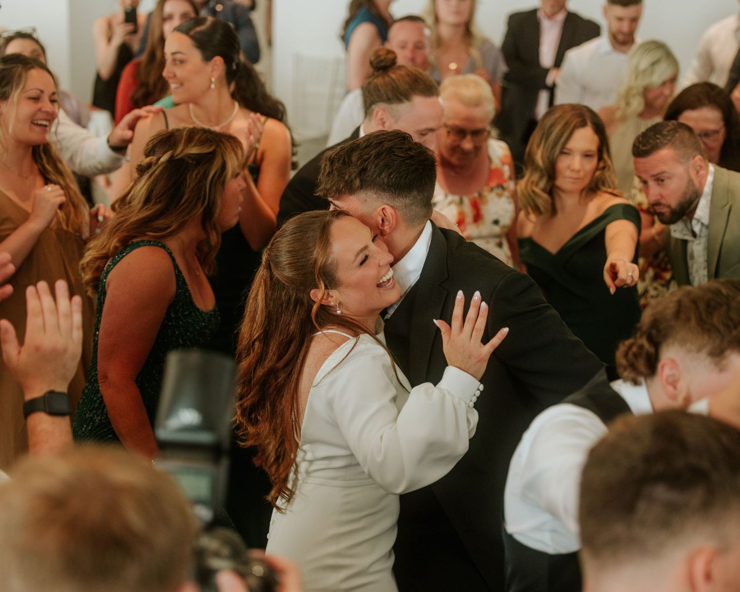 Bride and groom share a joyful embrace on the dance floor surrounded by celebrating wedding guests.