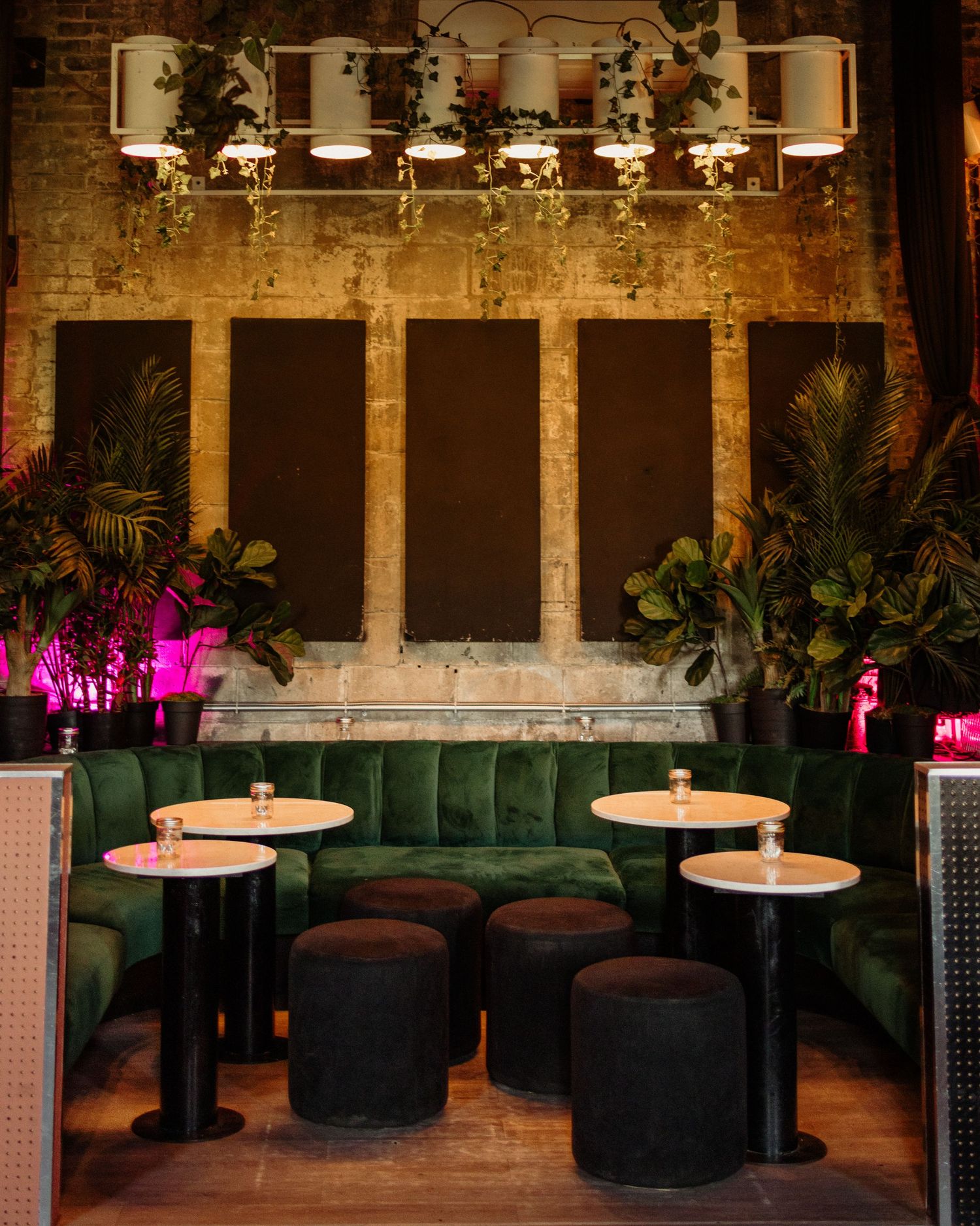 Lush green velvet booths with round tables, ottomans, tropical plants, and warm pendant lights in an upscale bar lounge.