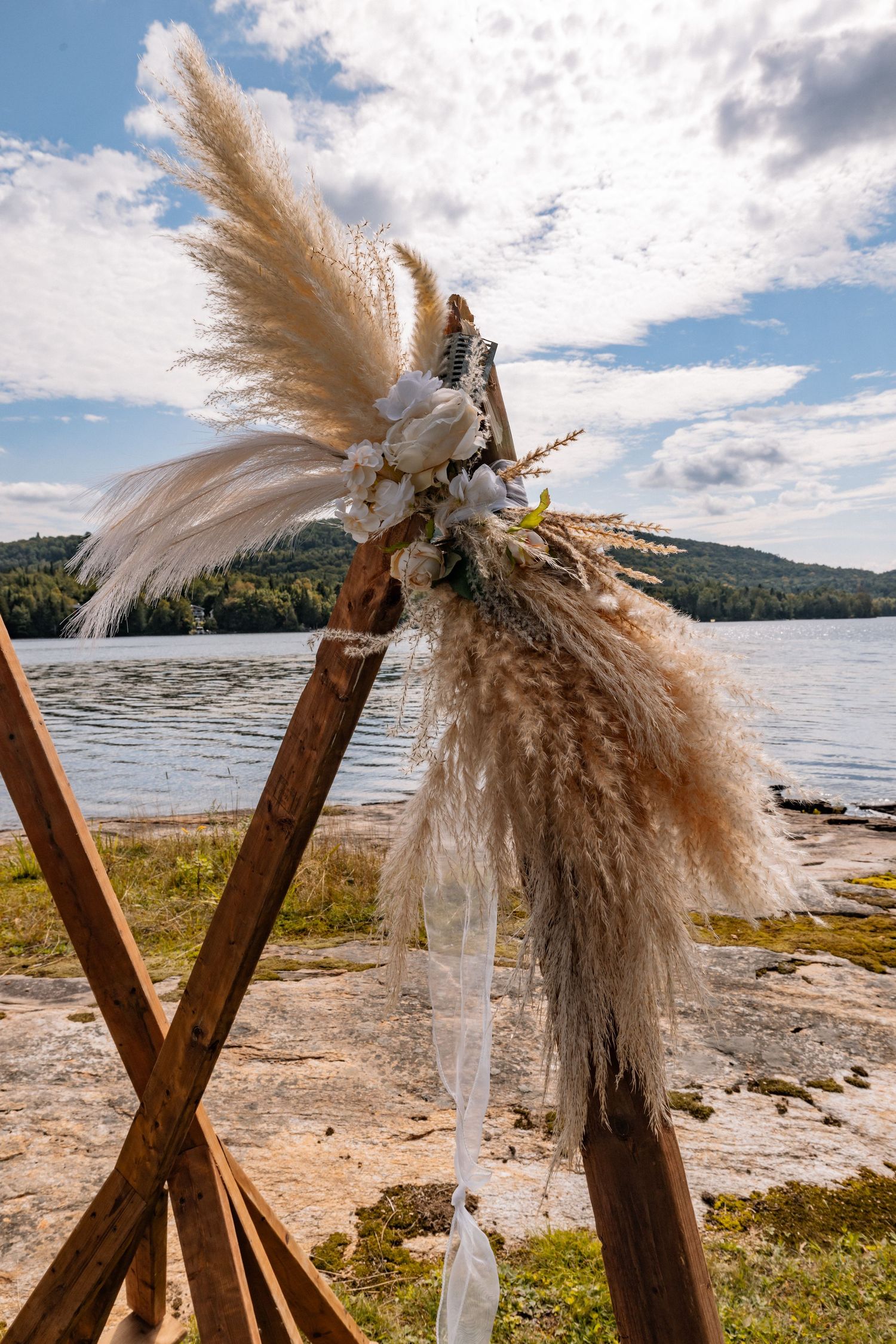 Boho wedding arch adorned with pampas grass and white feathers by a serene lakeside under a partly cloudy sky.