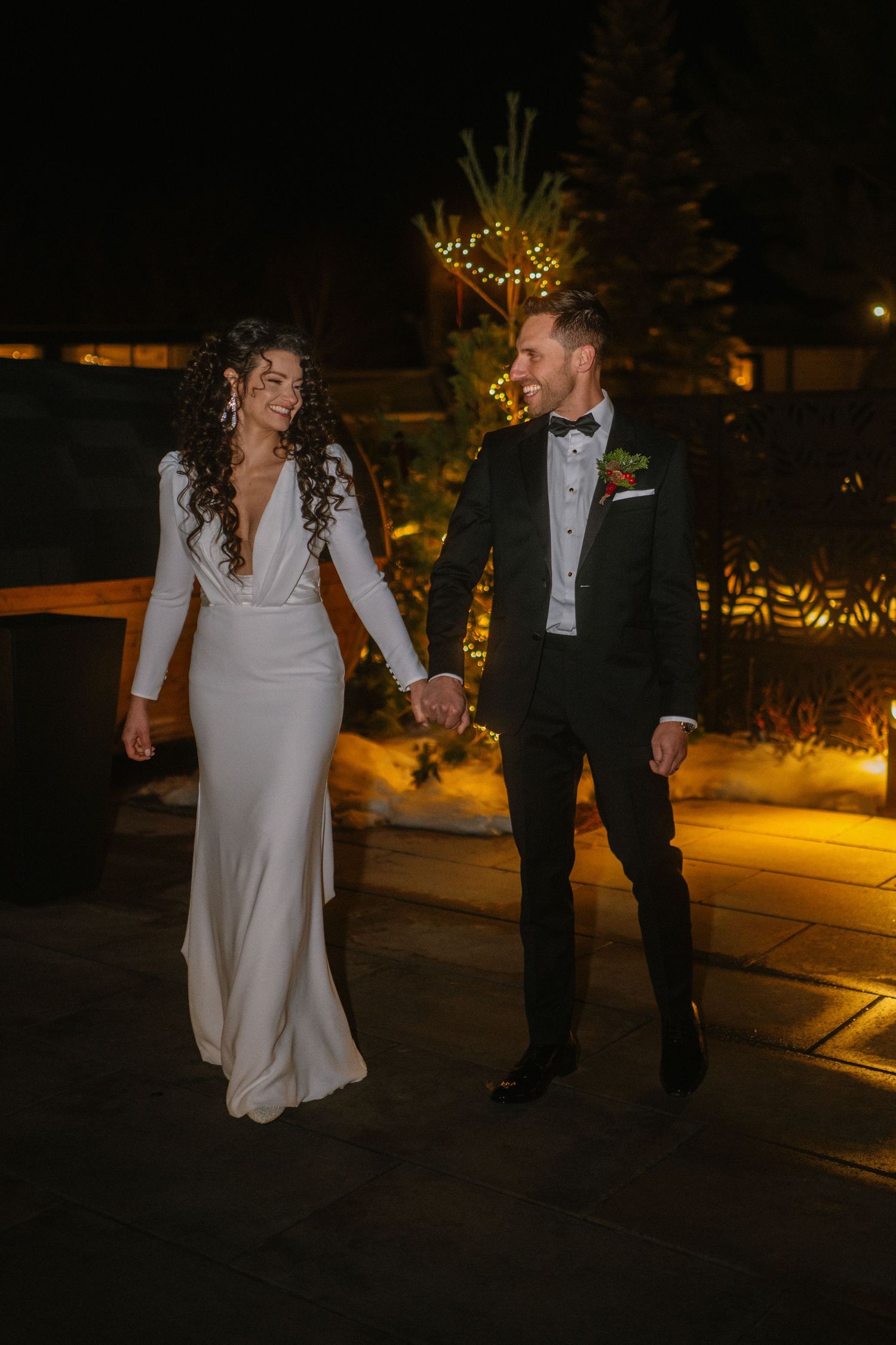 Bride and groom hold hands walking outdoors at night, she in a white gown, he in a black tuxedo with string lights behind.