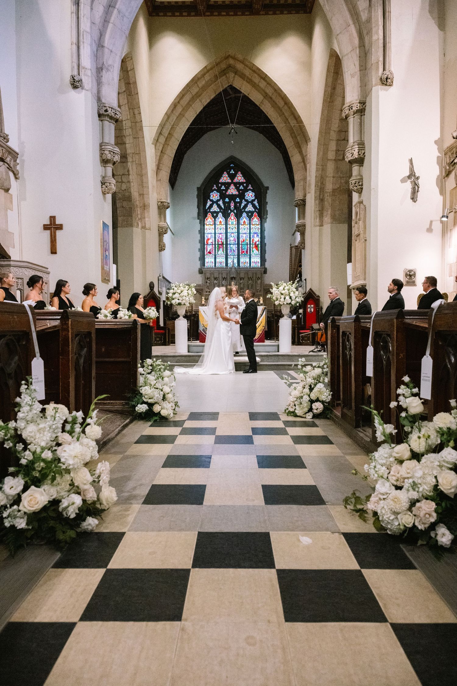 Bride and groom exchange vows at elegant church altar with white floral arrangements lining black and white checkered aisle.