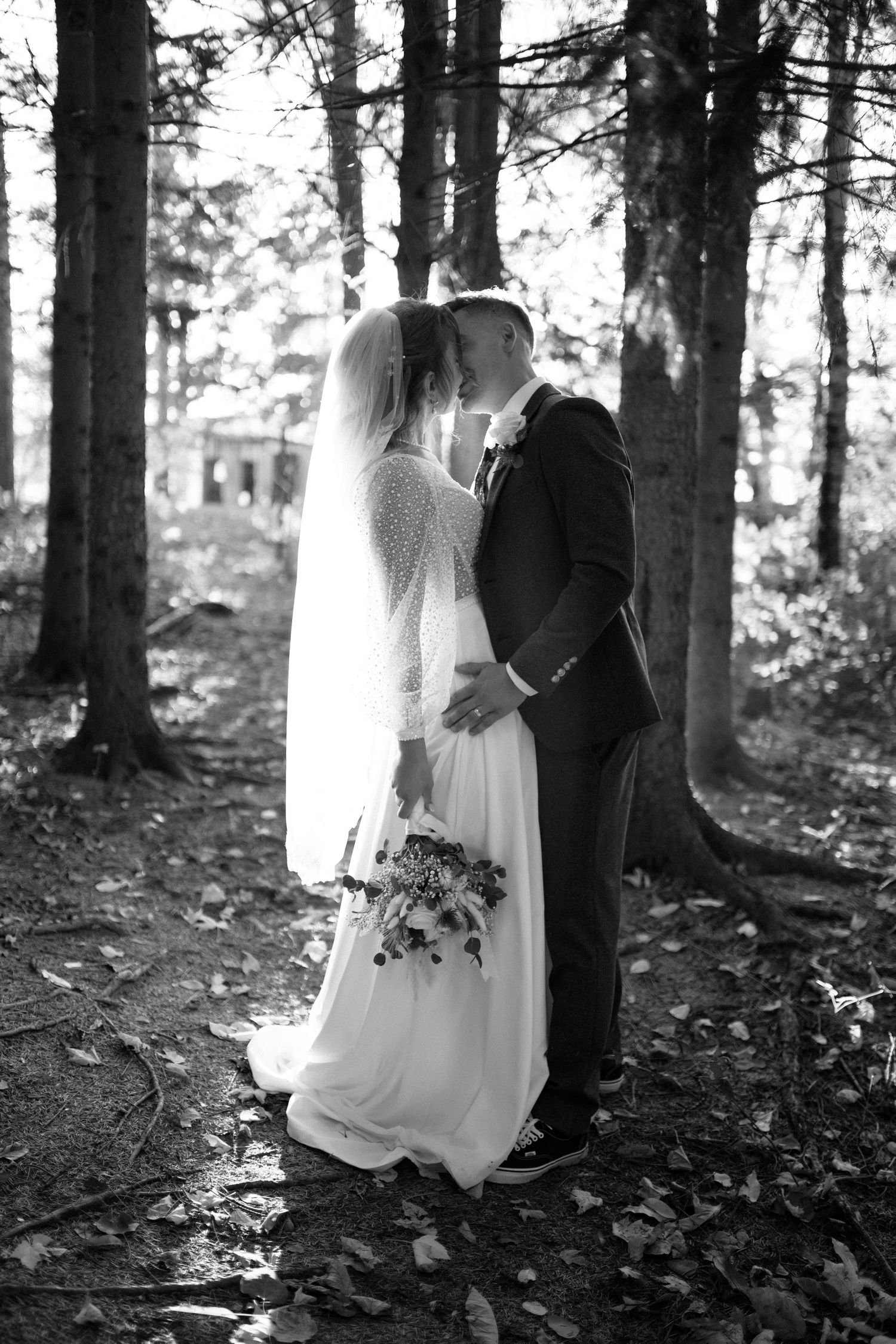 Black and white romantic photo of bride and groom kissing in a sunlit forest setting.