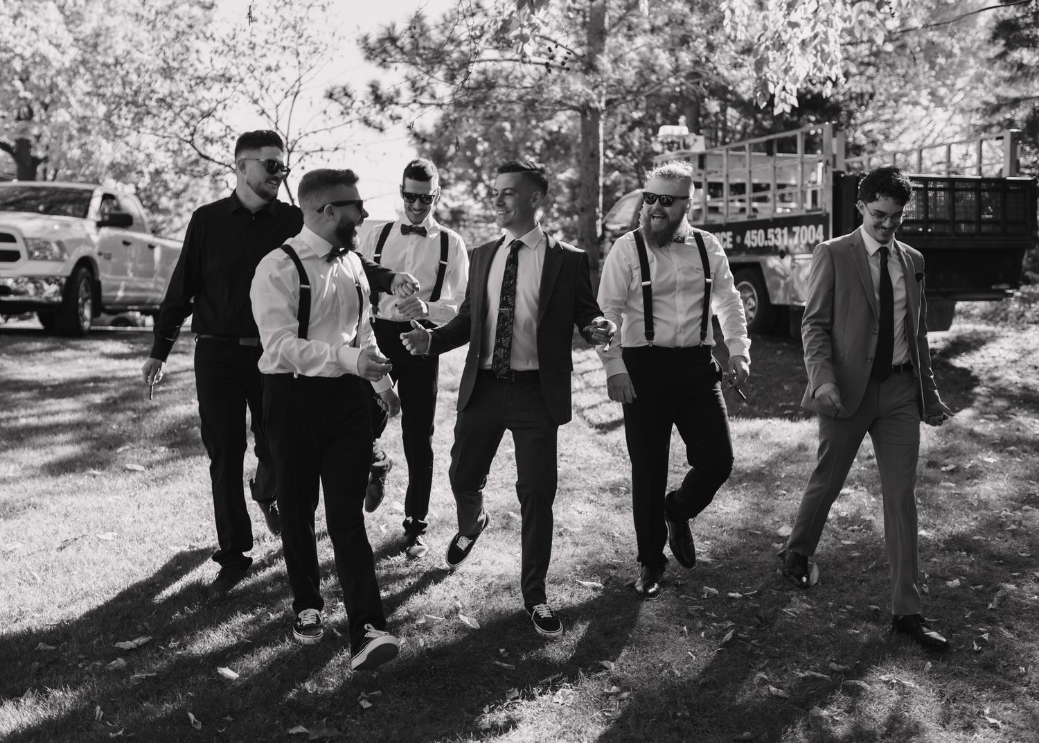 Groomsmen in suspenders and dress shirts walk outdoors in a black and white wedding photo.