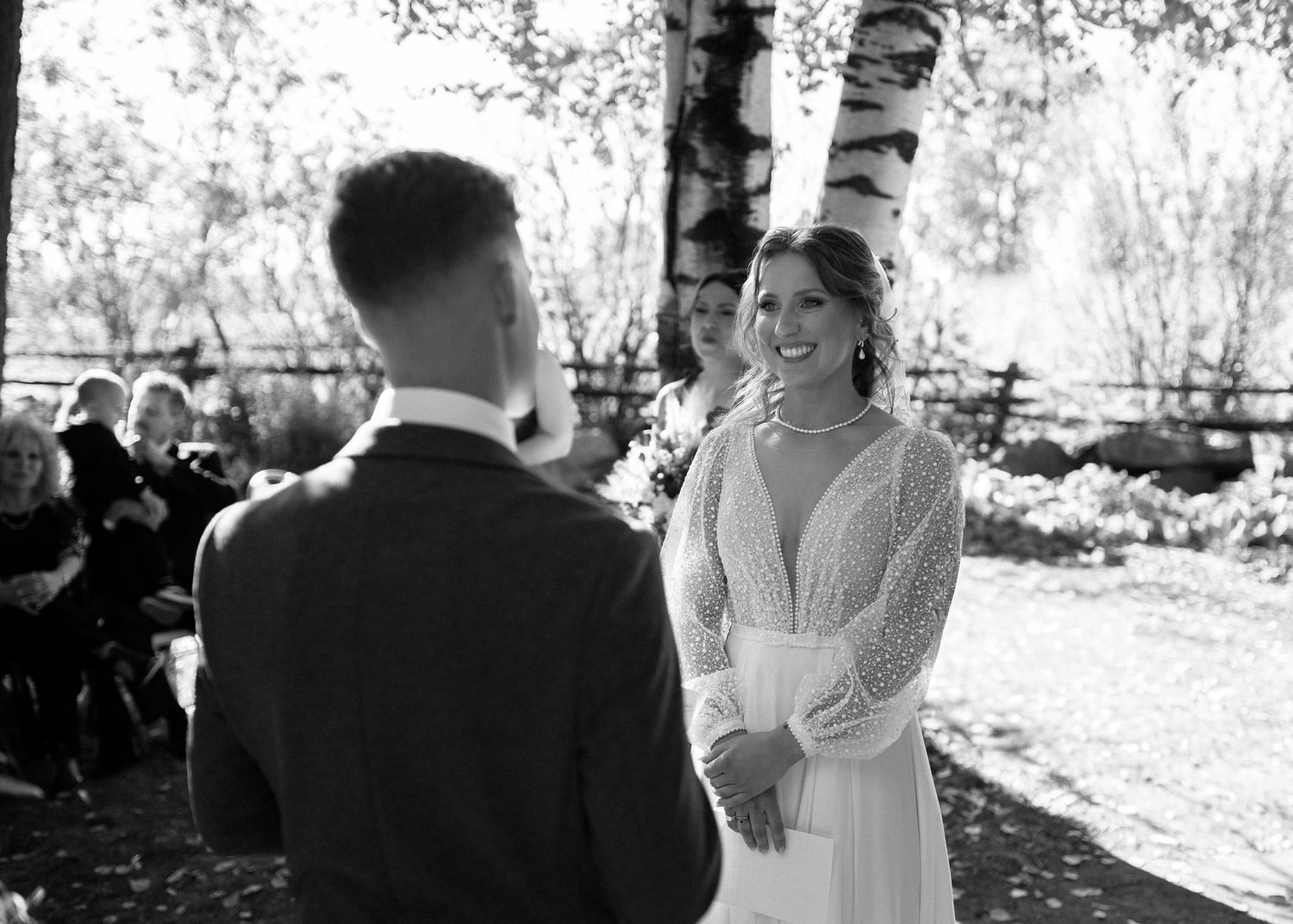 Black and white photo of smiling bride at outdoor wedding ceremony with groom and guests.