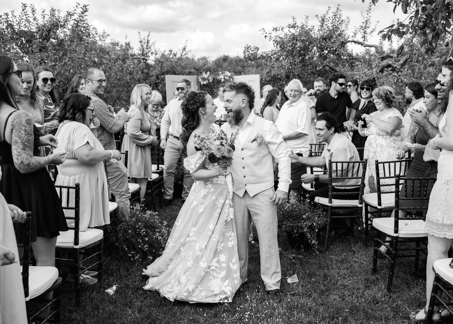 Black and white photo of newlyweds kissing at outdoor wedding ceremony aisle surrounded by guests.