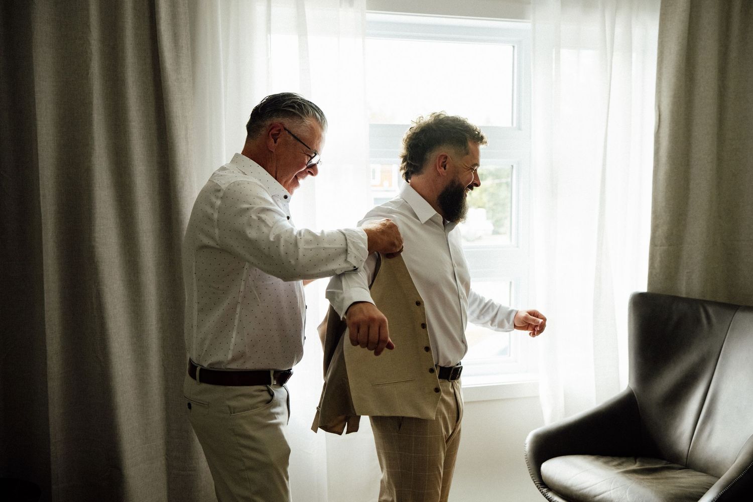 Two men in formal attire help each other with a suit jacket near a bright window, preparing for a special occasion.
