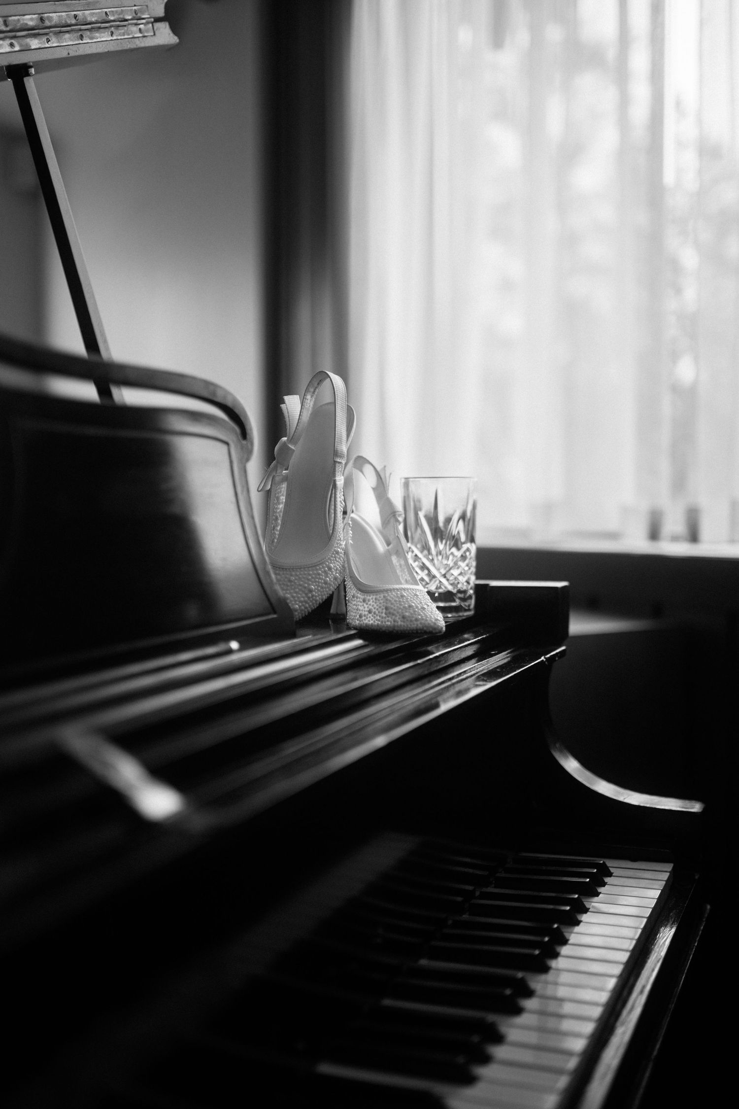 Black and white photo of bridal heels and crystal glass on a grand piano keys near a window.