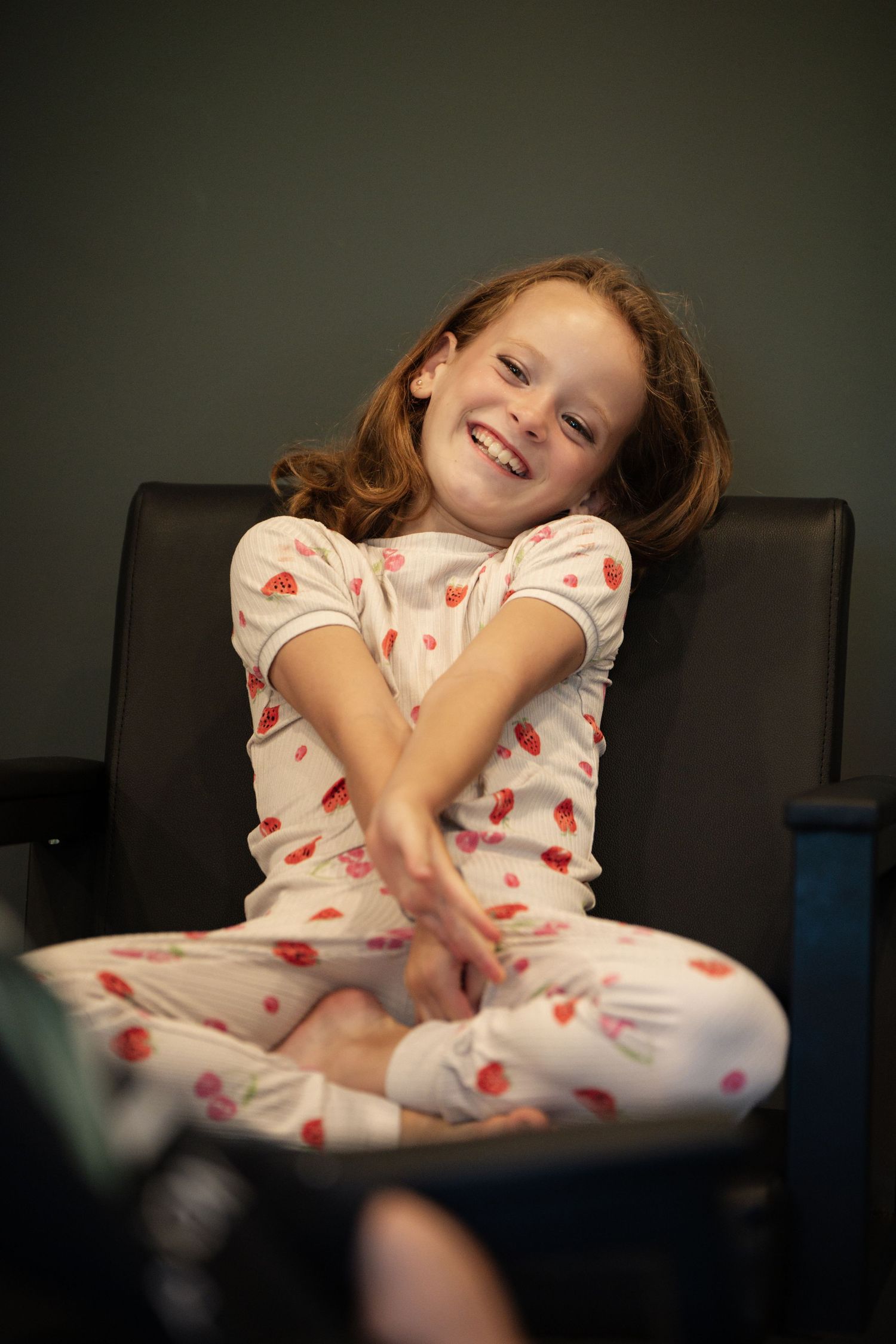 Smiling young girl in floral pajamas sitting cross-legged on a dark leather chair, reaching toward camera.