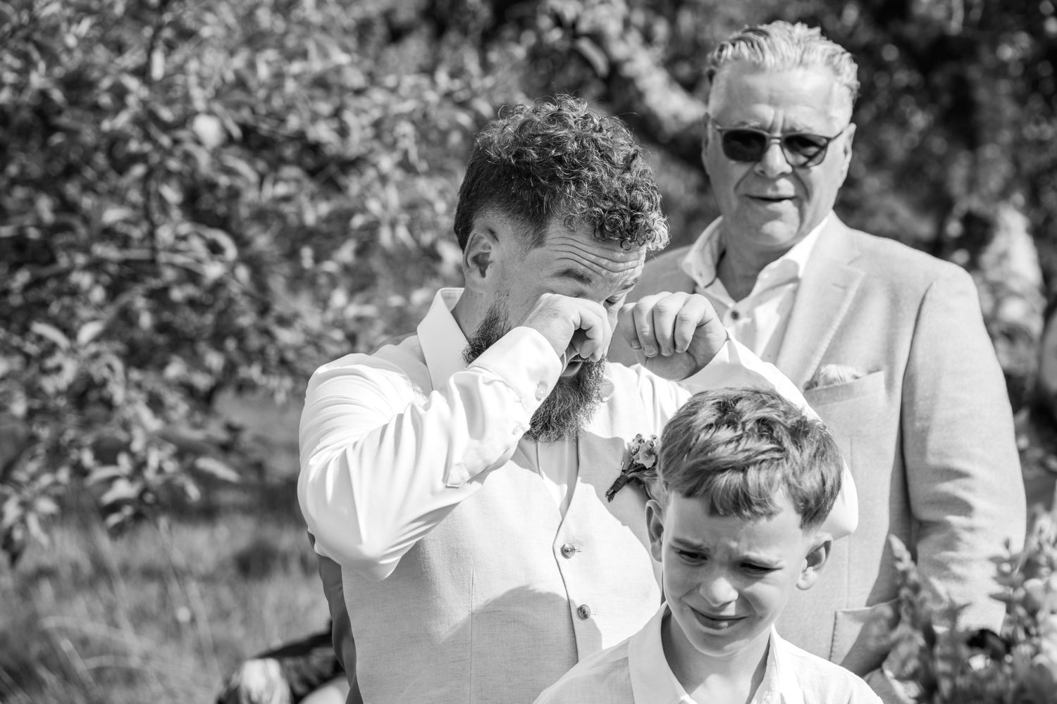 Emotional groom wipes tears during outdoor wedding ceremony while guests look on in black and white.