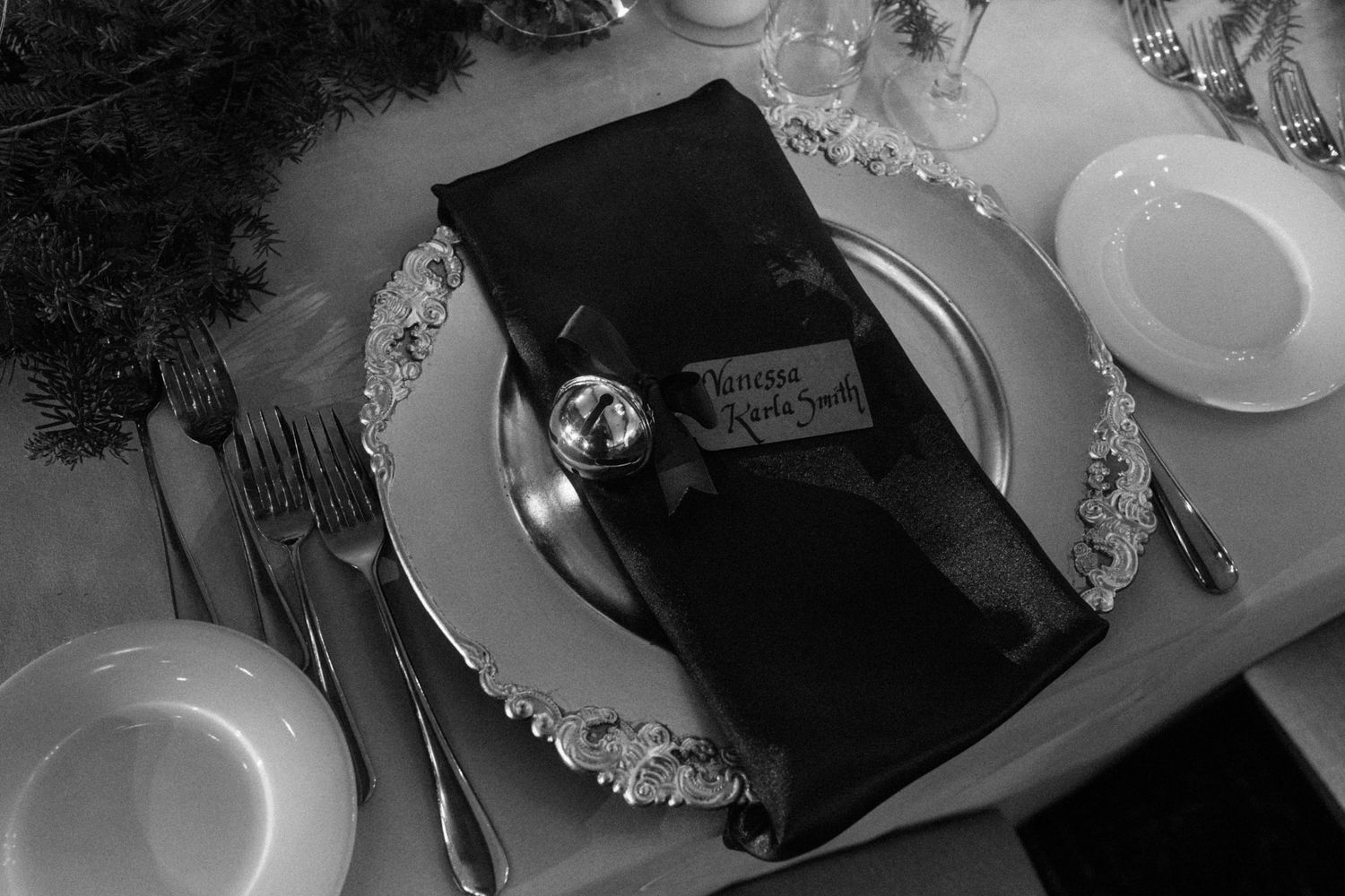 Black and white photo of an elegant formal place setting with silverware, plates, and a folded black napkin with a name card.