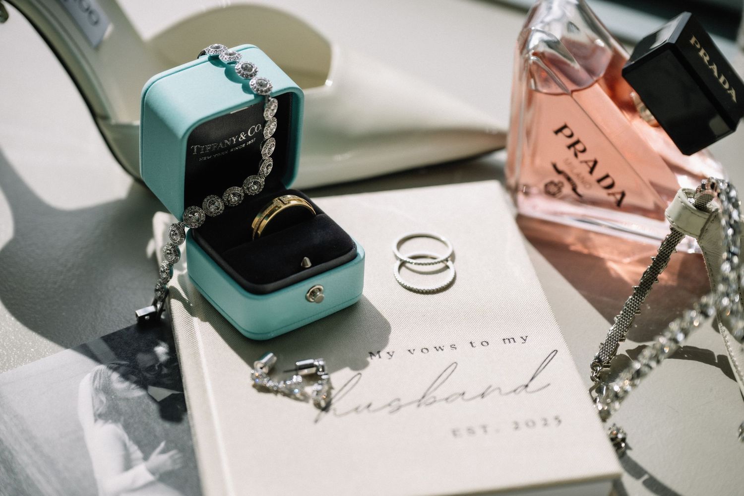 Elegant wedding flat lay with gold ring in teal box, Prada perfume, pearl bracelet, and vow book.