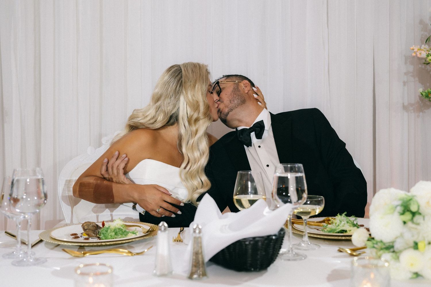 Newlywed couple sharing a romantic kiss at their elegant wedding reception table.
