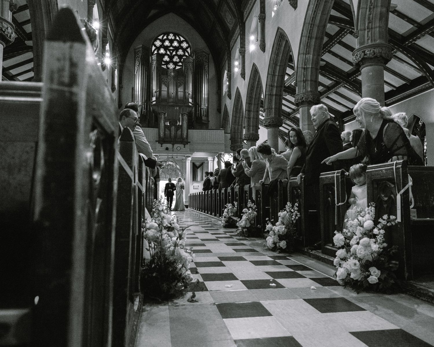 Black and white photo of wedding guests seated in ornate church pews lined with floral arrangements.