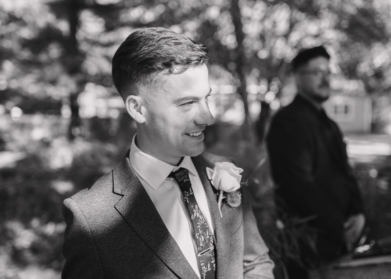 Black and white portrait of a smiling groom wearing a floral boutonniere and patterned tie outdoors.