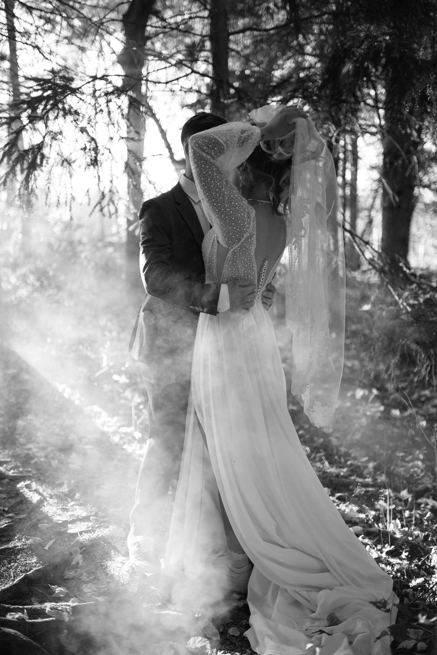 Black and white dramatic photo of a bride and groom kissing in a misty forest, surrounded by smoke and pine trees.