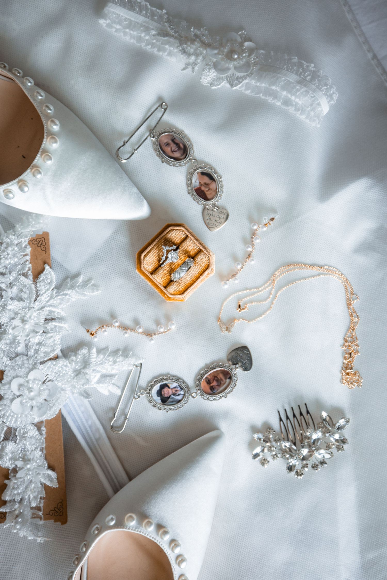 Bridal flat lay with pearl heels, lace garter, photo charms, gold ring box, crystal hair comb, and delicate necklace on white fabric.