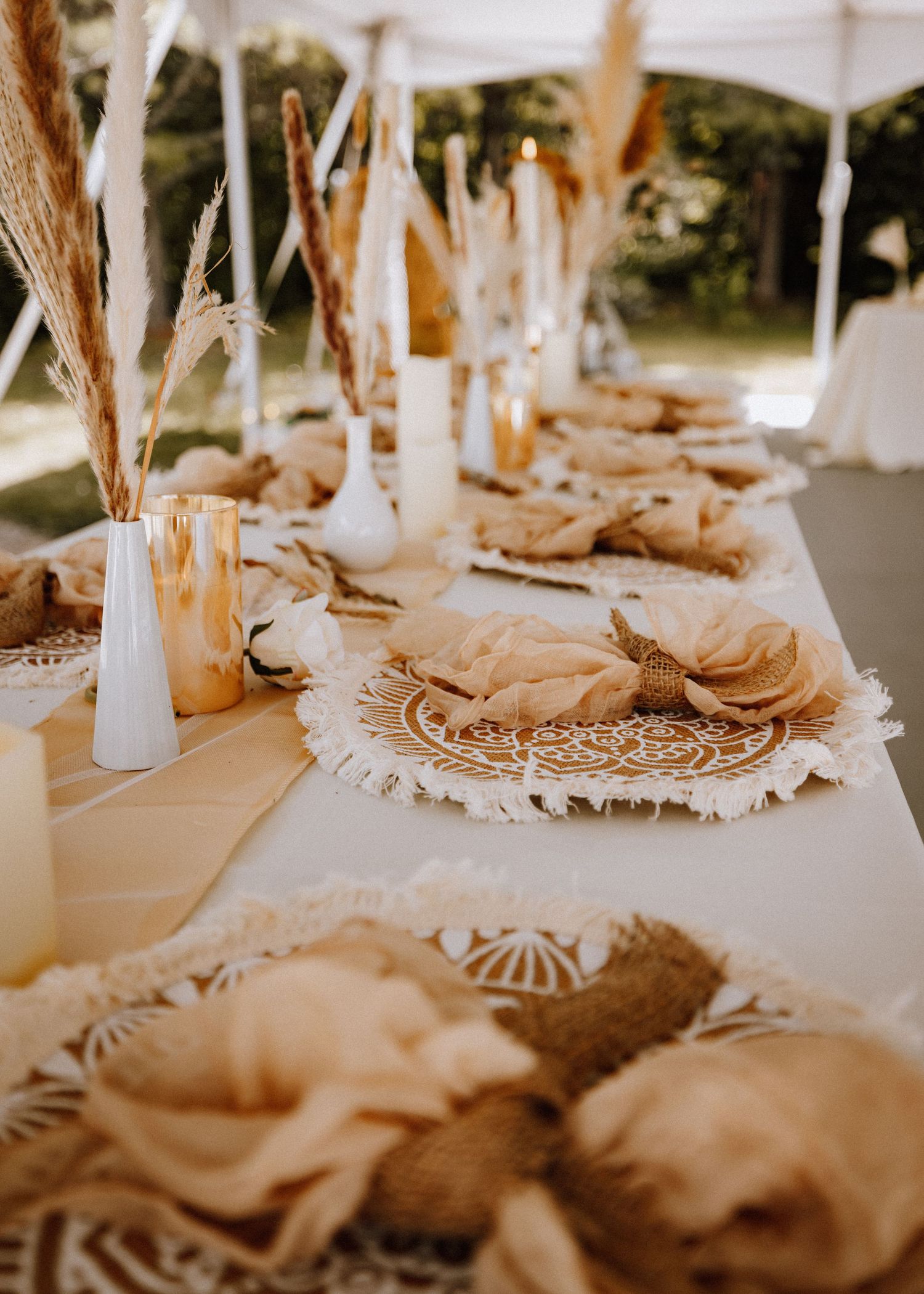 Boho wedding table setting with macrame placemats, pampas grass, white candles, and neutral linen napkins outdoors.
