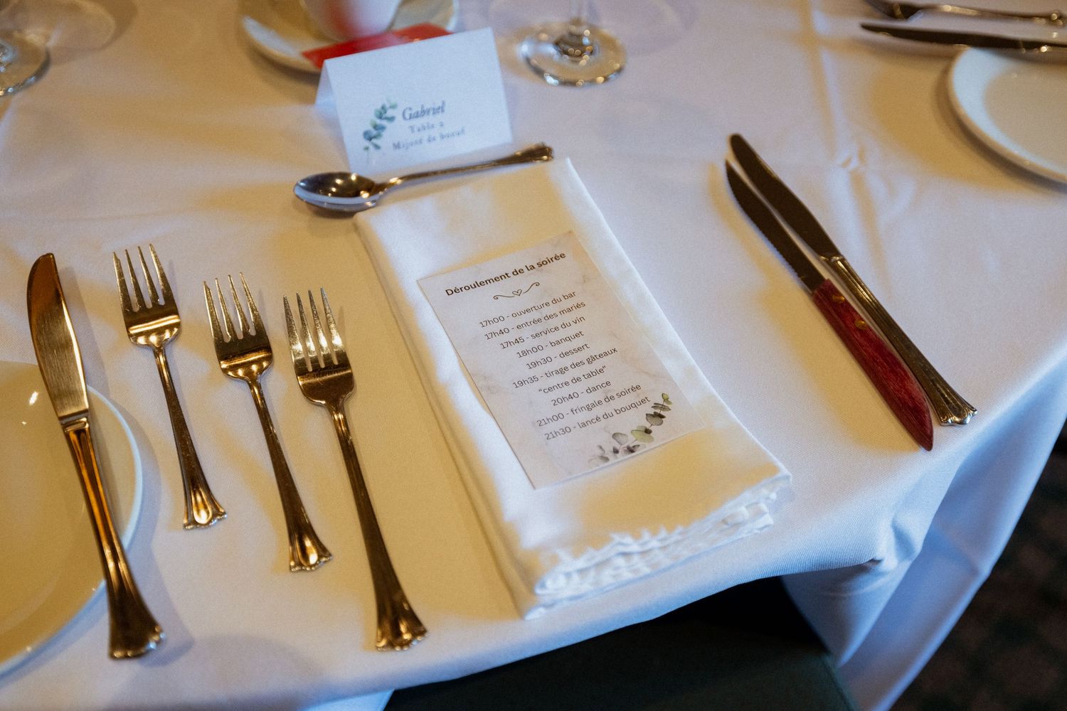 Elegant wedding reception place setting with gold silverware, white napkin, and printed menu on a white tablecloth.