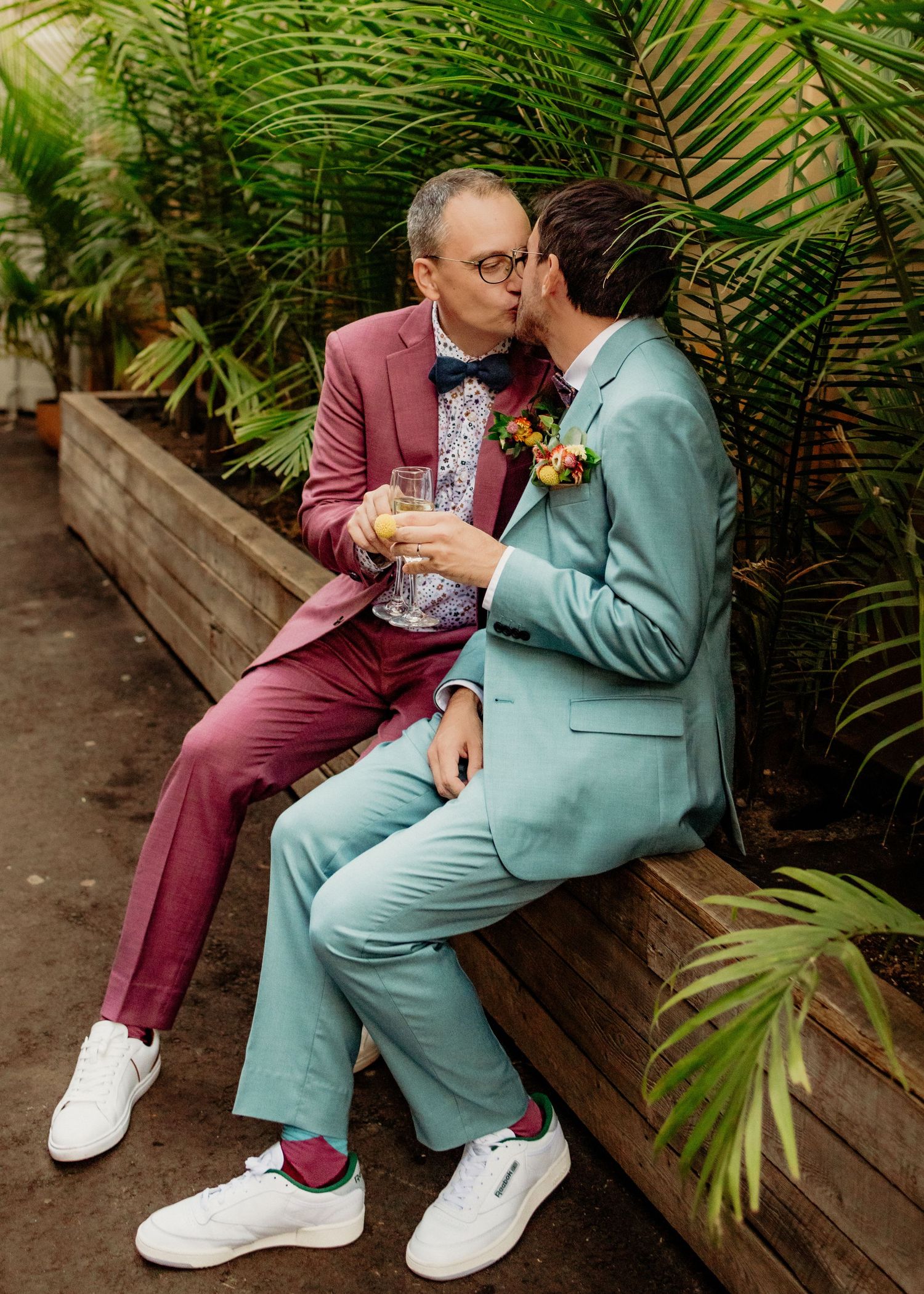 Two grooms in pink and teal suits share a kiss while seated outdoors among tropical plants.