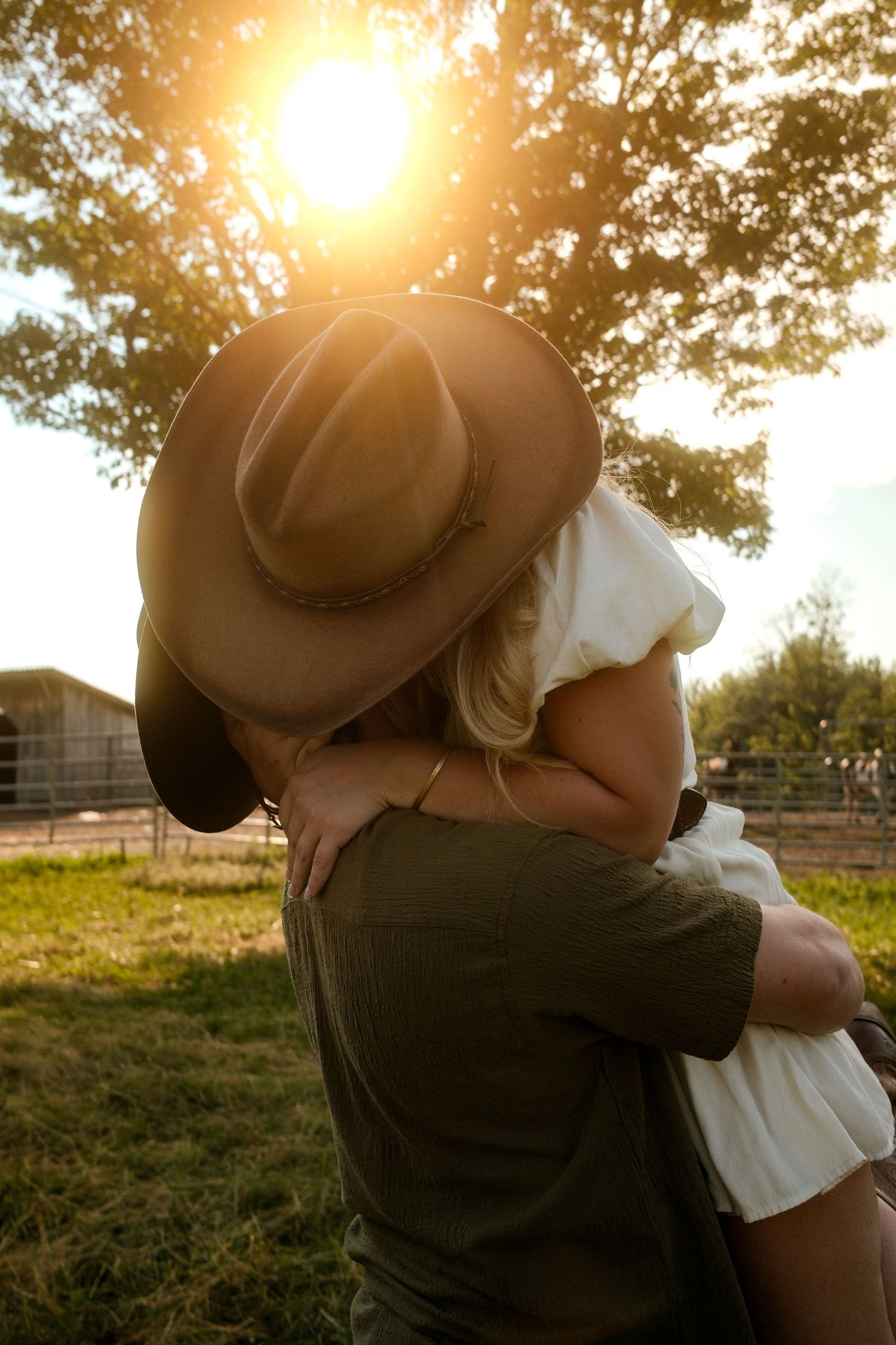 Couple embracing at golden sunset near farm, man wearing brown cowboy hat.