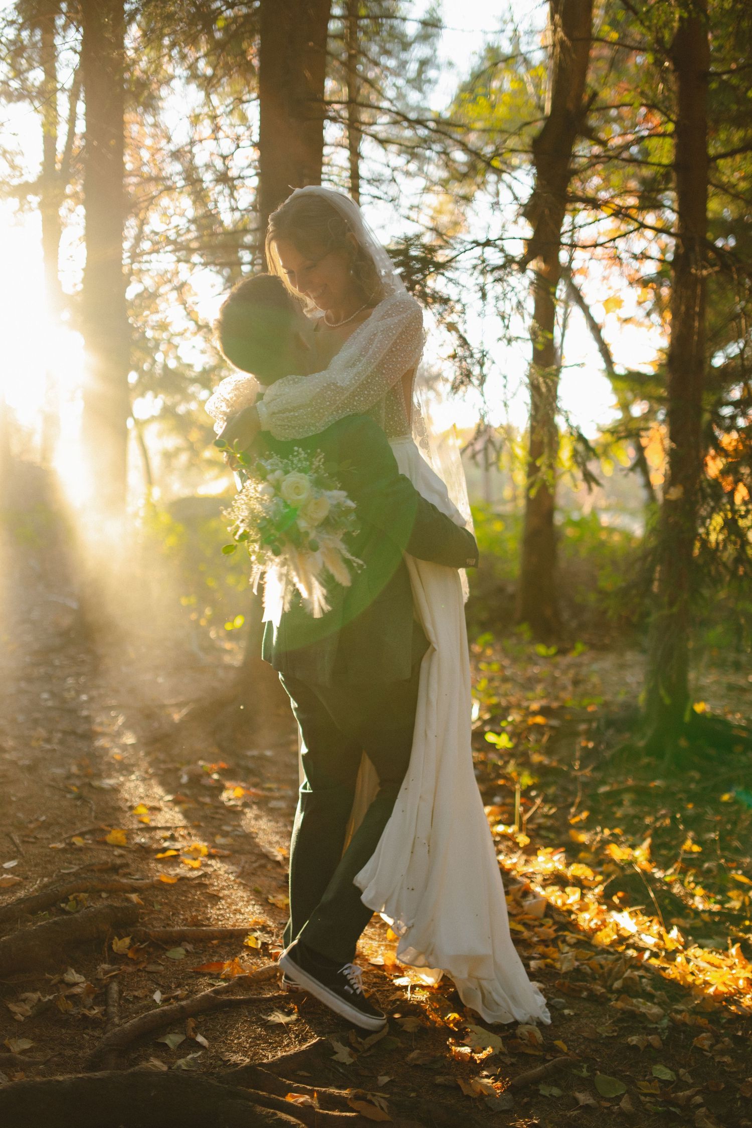 Bride and groom share a romantic embrace in sunlit autumn forest, she holds a white bouquet in a flowing dress.